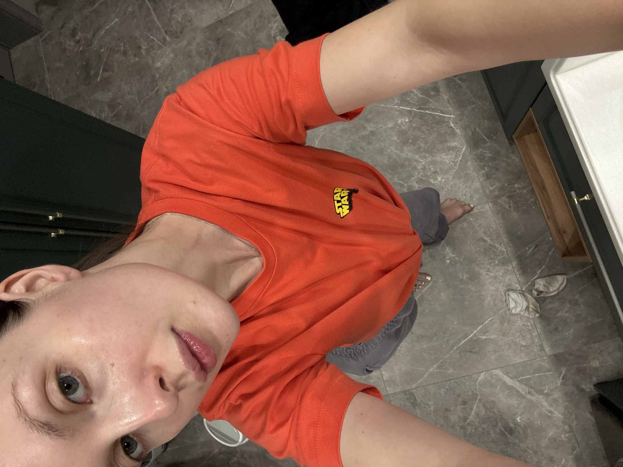 A person is taking a selfie in what appears to be a bathroom. The individual is wearing an orange t-shirt with a "Star Wars" logo on the chest and gray pants. The bathroom has gray marble floors and a dark green cabinet. There is a white countertop and a drawer with a wooden interior. The location of the photo is not definitively identifiable as Tomilino, Russia without additional information.