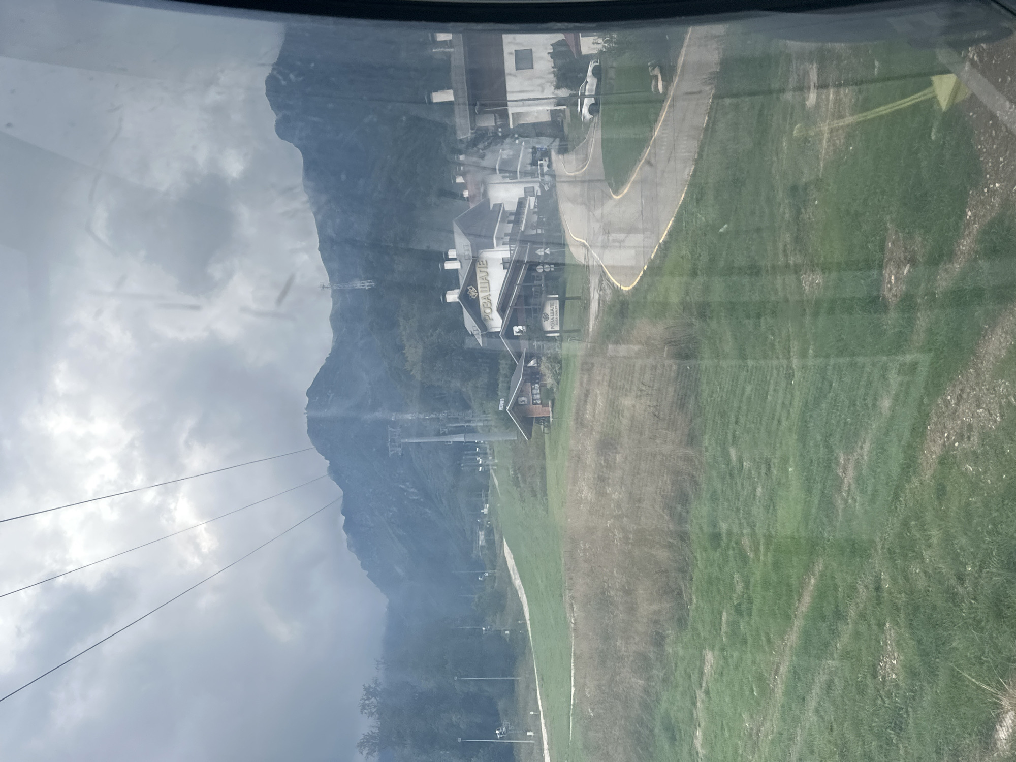 The image captures a view from what appears to be a ski lift or cable car, looking down on a mountain resort area. In the foreground, a grassy slope with dry patches is visible, and further down, a paved road winds through a cluster of buildings. These buildings, constructed in a traditional Alpine style with pitched roofs, are predominantly white with dark trim. One building prominently displays the sign "РОЗА ШАЛЕ" in large letters. Several cars, including a white luxury sedan, are parked along the road or in front of the buildings.
In the middle ground, a ski slope is visible, marked by poles and what looks like a ski lift apparatus. Trees and more ski lift towers are scattered across the landscape, leading up to a range of imposing, mist-shrouded mountains in the background. The sky is overcast with dark, heavy clouds, suggesting a potentially stormy or cloudy day. The overall scene depicts a resort town nestled in a mountainous region under a dramatic sky. The location is identified as Estosadok, Russia, a popular mountain resort.
