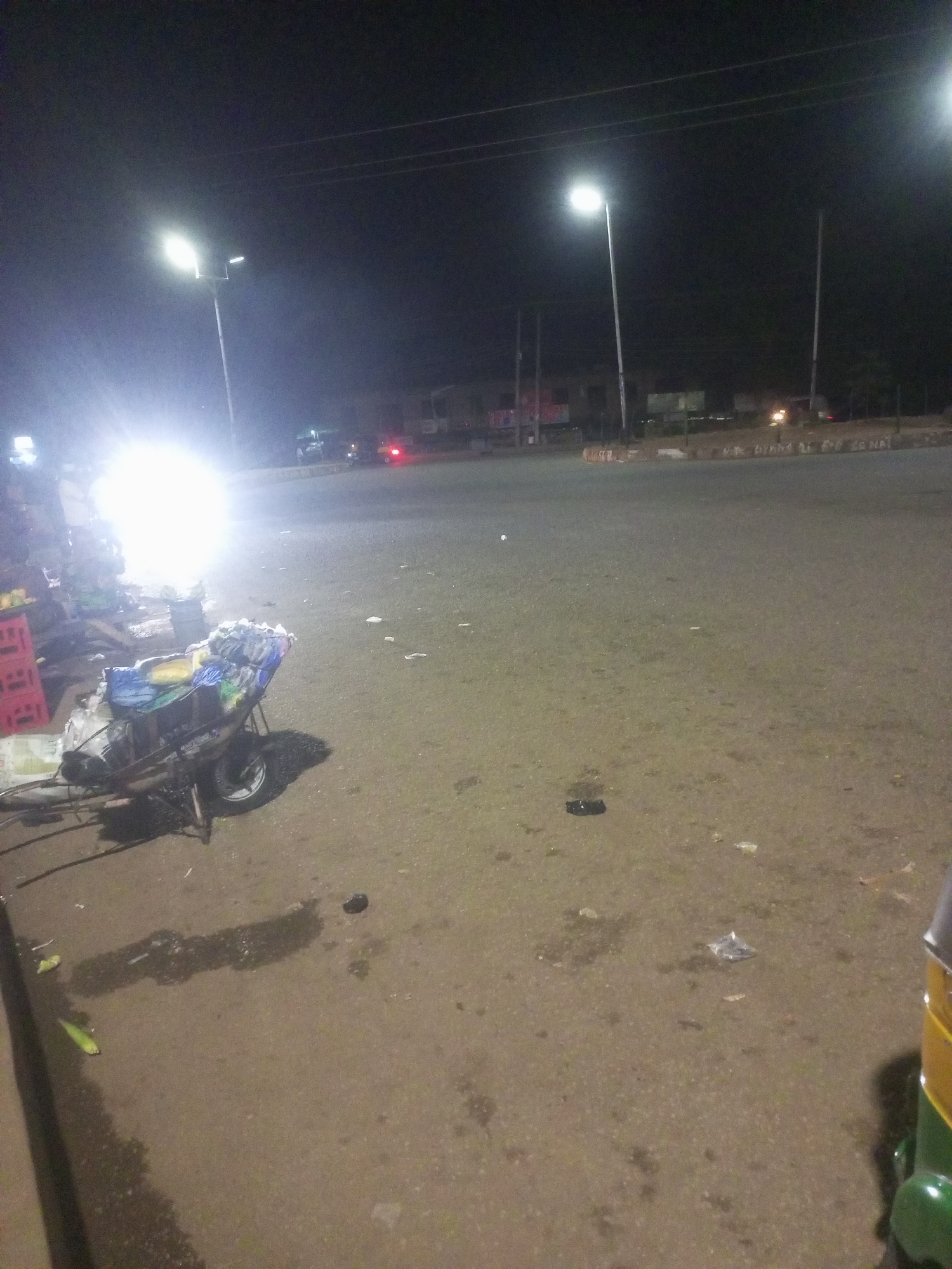 The image depicts a nighttime scene on a paved road or intersection in Bukuru, Nigeria. The sky is dark, indicating a late hour, and the area is primarily illuminated by artificial light sources.
On the left side of the frame, an intensely bright light, possibly from a vehicle or a powerful lamp, creates significant lens flare and overexposure. Near this light, a two-wheeled cart or wheelbarrow filled with various items is visible, along with stacked red plastic crates. The bright light casts long shadows of these objects across the ground.
Several tall street light poles with modern, bright luminaires stand along the road, providing additional illumination. In the distant background, faint outlines of buildings can be discerned. Overhead wires are subtly visible against the dark sky.
The road surface appears to be asphalt or concrete and is scattered with litter, including plastic bags and other small debris. On the far right, a portion of a green and yellow vehicle, likely an auto-rickshaw, is partially visible. A low curb or barrier in the background has some faint, partially obscured white text, which appears to be a political slogan, possibly including "MR. D... FOR SENATE."
No people are visible in the frame, and the scene appears quiet and still.