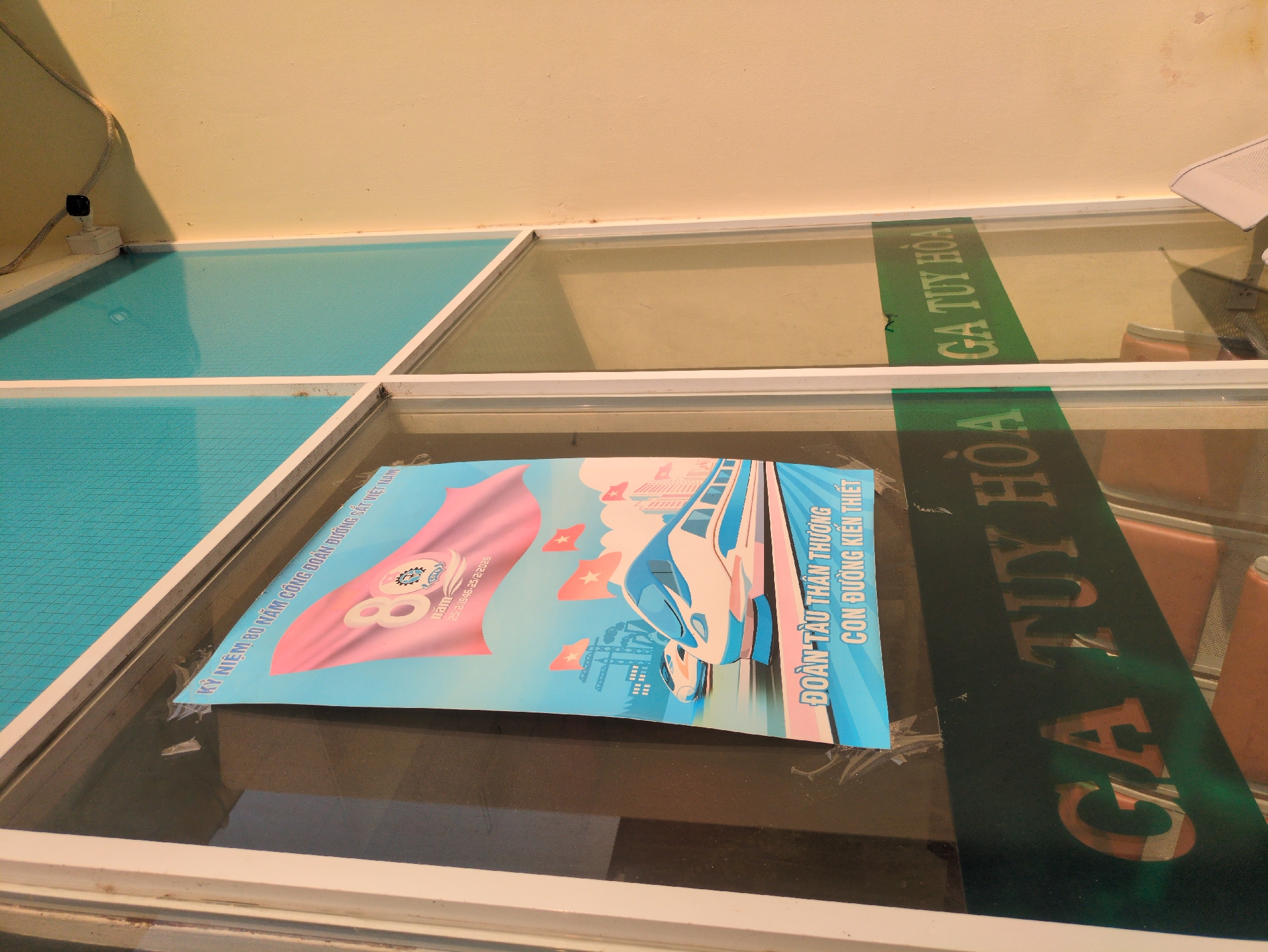 The image shows a poster commemorating the 80th anniversary of the Vietnam Railway Workers' Union (1946-2026), placed under a glass table. The poster features an image of a train and text that includes the phrases "ĐOÀN TÀU THÂN THƯƠNG - CON ĐƯỜNG KIẾN THIẾT" (Beloved Train - Building the Road). In a neighboring section of the glass table, the text "GA TUY HÒA" is visible, indicating that this scene is located in Tuy Hòa, Vietnam.