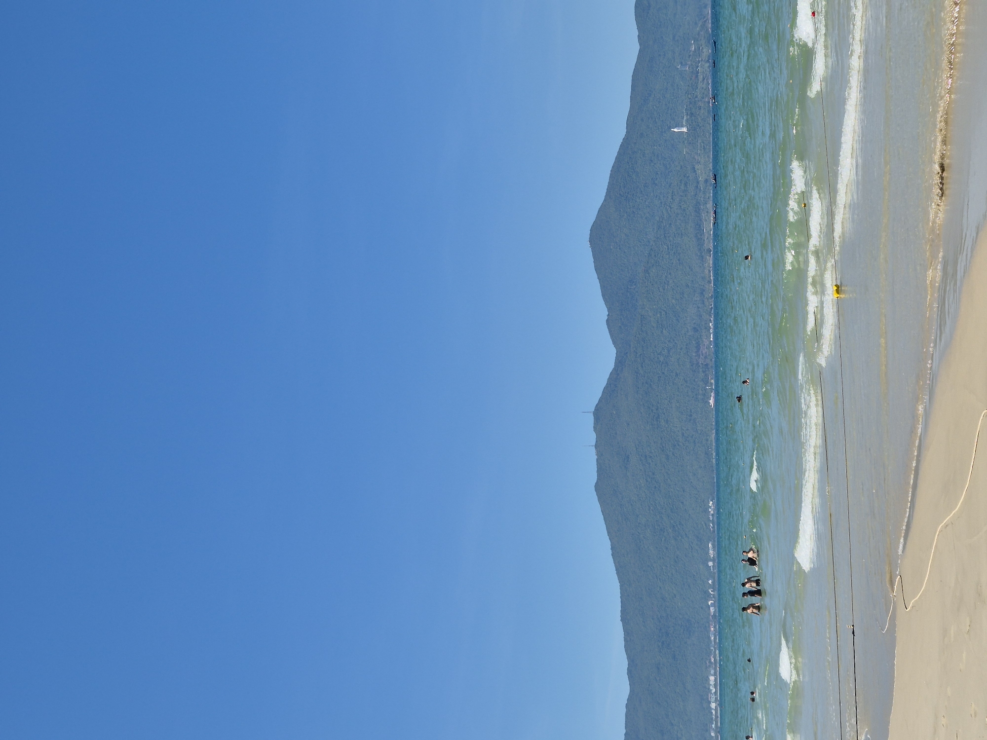 This is a landscape photograph taken at a beach in Da Nang, Vietnam. The image is oriented vertically. The left side of the frame is dominated by a vast expanse of clear blue sky. On the right side, a majestic, lush green mountain range slopes down to meet the ocean. The sea is a vibrant turquoise, with gentle waves breaking along the shore. The beach itself is sandy and appears to be quite wide.
Several people are visible in the water, swimming and enjoying the ocean. Some are close to the shore, while others are further out. In the water, there are buoys and ropes, possibly marking swimming areas or separating different zones. One person is using a yellow flotation device. The sun is high in the sky, suggesting it is midday. The weather appears to be clear and sunny, typical for a beach day. In the distance, along the coastline at the base of the mountains, there are hints of buildings, suggesting a populated area or resort. The overall mood of the image is peaceful and relaxed, conveying a sense of summer leisure and enjoyment. There is no visible text in the image.