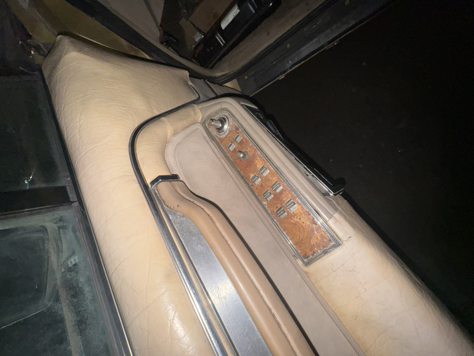 The scene captures the interior of a vehicle, specifically a front door panel, located in Zhabkino, Russia. The door panel is upholstered in worn, light-tan padded material, exhibiting creases and dirt accumulation. A horizontal chrome trim piece is present above a recessed armrest section, which includes a matching tan door pull handle. Below the armrest, a control module with a distressed faux woodgrain finish houses four silver-colored rectangular power window switches, arranged in two columns of two. To the right of these switches are two circular controls, one larger and one smaller, for unspecified functions. The lower portion of the image shows a dark floorboard inside the vehicle and dark ground outside the open door. Indistinct dark objects are visible on the vehicle's floorboard to the far right. A dirty vehicle window is partially visible in the upper left. No individuals are present.