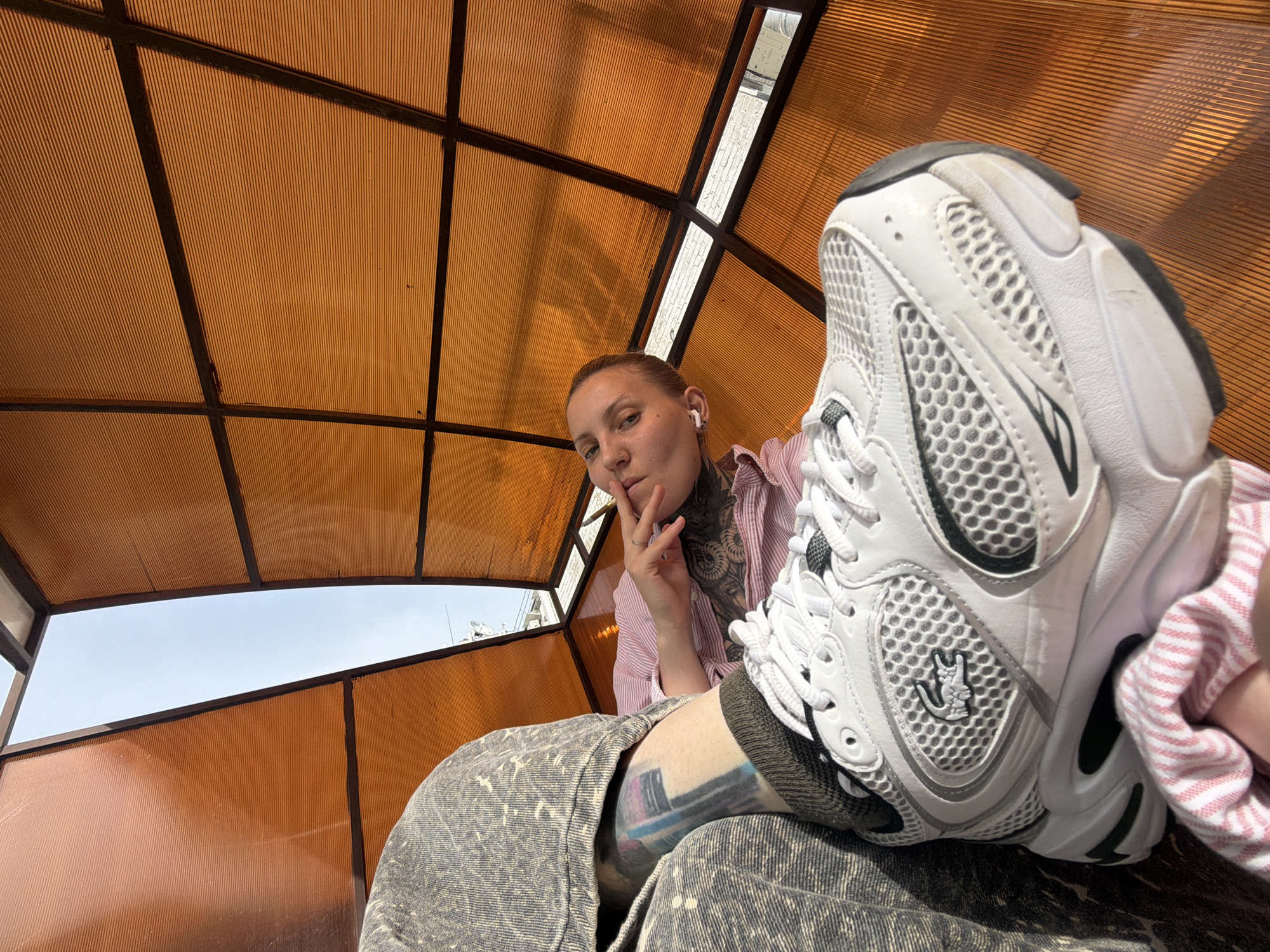 A low-angle shot depicts a person seated under a brown, gridded structure possibly in Chaikovsky, Russia. The person has short hair, a pierced nose, and appears to be wearing wireless earbuds. Their left hand is raised near their face, with a finger pointing up. They are dressed in a pink and white striped shirt, with tattoos visible on their neck and arm. The person's legs are covered in denim patterned trousers. A white athletic shoe with black trim and a logo on the side is prominently displayed, resting on their leg. Behind the structure, a glimpse of the sky and some buildings can be seen. The structure is made of orange colored panels and metal beams.