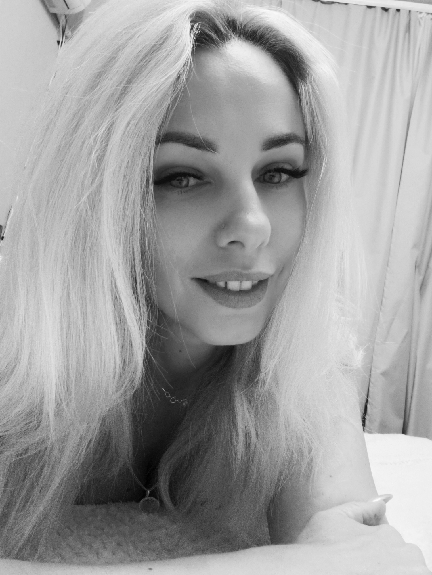 A grayscale portrait shows a woman with long, light-colored hair smiling slightly at the camera. She has noticeable eyelashes and is wearing a necklace. The background includes what appears to be a curtain or sheet of fabric. The location of the image cannot be determined to be in Sevastopol, Ukraine.