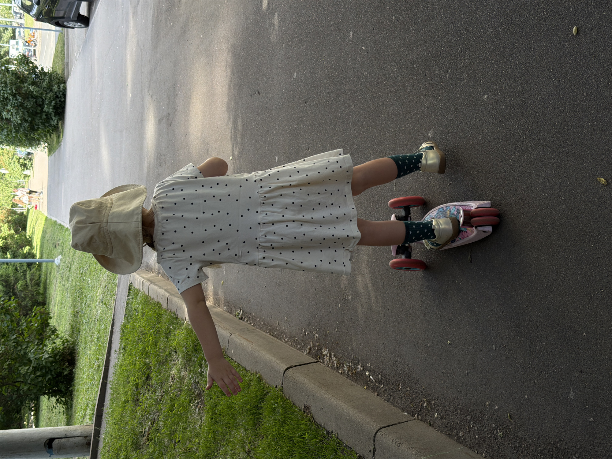 The image depicts a young child from a rear perspective, positioned on a pink and multi-colored three-wheeled scooter on a dark asphalt path in Moscow, Russia. The child wears a light tan bucket hat, an off-white short-sleeved dress with small black polka dots, dark green polka-dotted socks, and light gold-colored sandals. The child's left arm is extended horizontally outwards, palm open. To the left, a concrete curb separates the path from a grassy area with varied green foliage and a concrete utility pole. Fine, light-colored debris is accumulated along the curb's edge. In the background, two adult figures walk along a distant section of the path, and a dark-colored vehicle is partially visible on an adjacent paved surface to the right. The scene is illuminated by daylight.