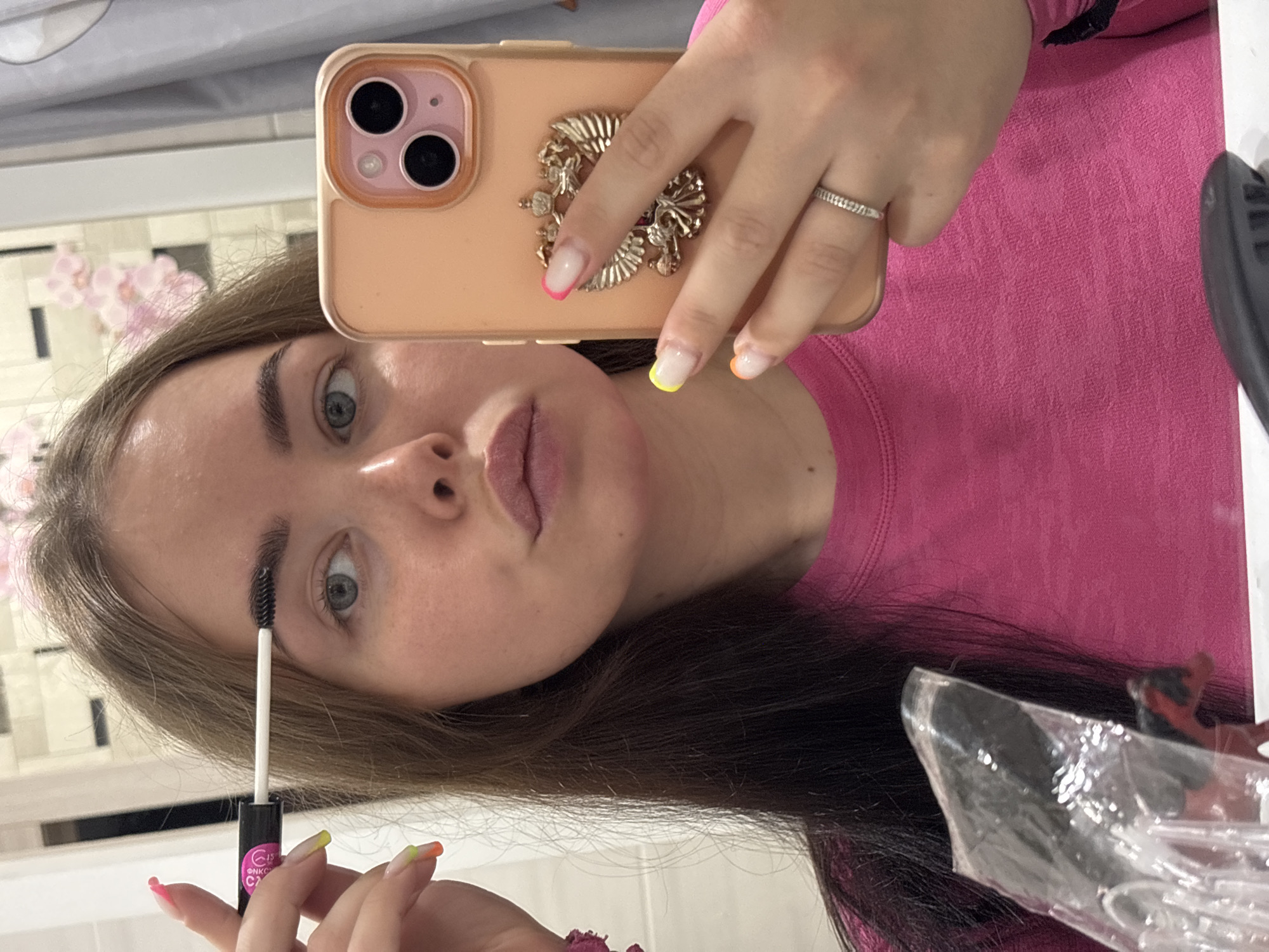A young woman is captured in a mirror selfie while applying makeup indoors. She has long brown hair and is wearing a bright pink long-sleeved top. Her attention is on her reflection as she grooms her eyebrows with a brush from a black tube, which bears Cyrillic text "ФНКС" (FNKS), numbers "15" and "С2", and a logo.
Her right hand holds a light peach or orange-cased iPhone, which has an elaborate gold-colored crest or emblem as a phone grip. A textured silver-colored ring is visible on her right ring finger. Her nails are styled with a French manicure featuring vibrant tips in various colors: pink, yellow, orange, and green. She has light-colored eyes and appears to be wearing a light lip product.
The background, visible through the mirror, suggests an indoor setting, possibly a bathroom or vanity area, with bright lighting, some light-colored tiled surfaces, and subtle pink floral decorations. The image is taken at a 90-degree rotation, with the subject's head tilted to the right. The activity takes place in Izhevsk, Russia.