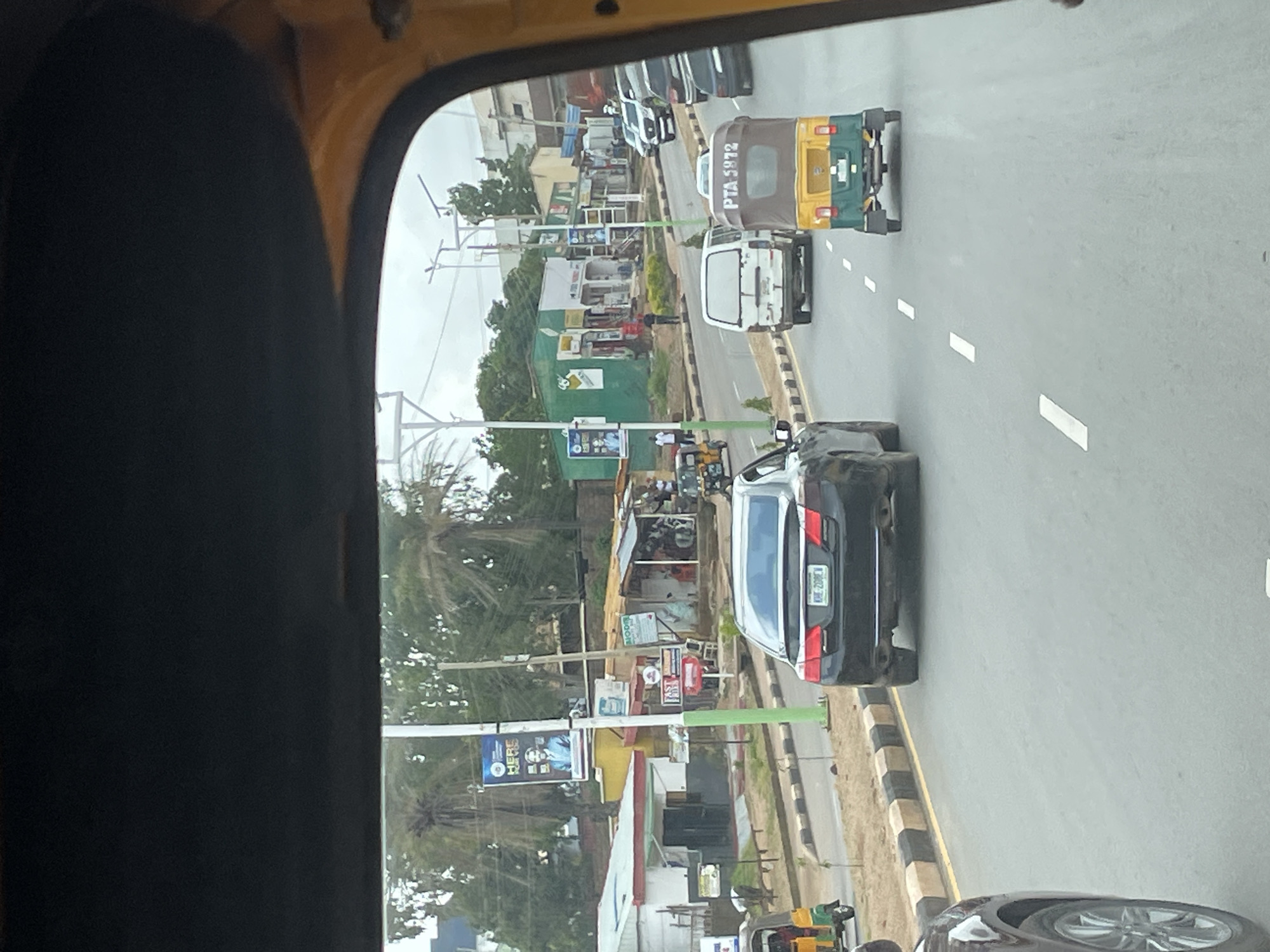 This is a photograph taken from inside a vehicle, likely an auto-rickshaw, looking out at a street scene in Jos, Nigeria. The perspective is from the driver's or front passenger's viewpoint, with the dark interior of the vehicle dominating the left side of the frame.
The scene depicts a moderately busy street with several vehicles, including a yellow and green auto-rickshaw with the license plate "PTA 5972," a white van, and several cars. The vehicles are traveling in the same direction, suggesting a flow of traffic. On the right side of the street, there are buildings that appear to be shops or businesses, with awnings and signs visible. Some of these buildings have a more rustic or commercial appearance, with a green building standing out.
Along the side of the road, there are various signs. One prominent sign has text that is partially obscured but appears to be an advertisement or announcement, with a picture of a person's face. Another sign is red and white, and the text "FAST" is clearly visible. There are also utility poles with wires, indicating the presence of infrastructure.
The time of day appears to be daytime, likely late morning or afternoon, given the bright ambient light and the shadows cast. The weather seems to be overcast or hazy, as the sky is not clearly visible and the light is diffused. The overall impression is of a bustling urban or semi-urban environment in Nigeria. There are no explicit emotional cues visible, and the activity is simply the ongoing movement of traffic and daily life on the street.