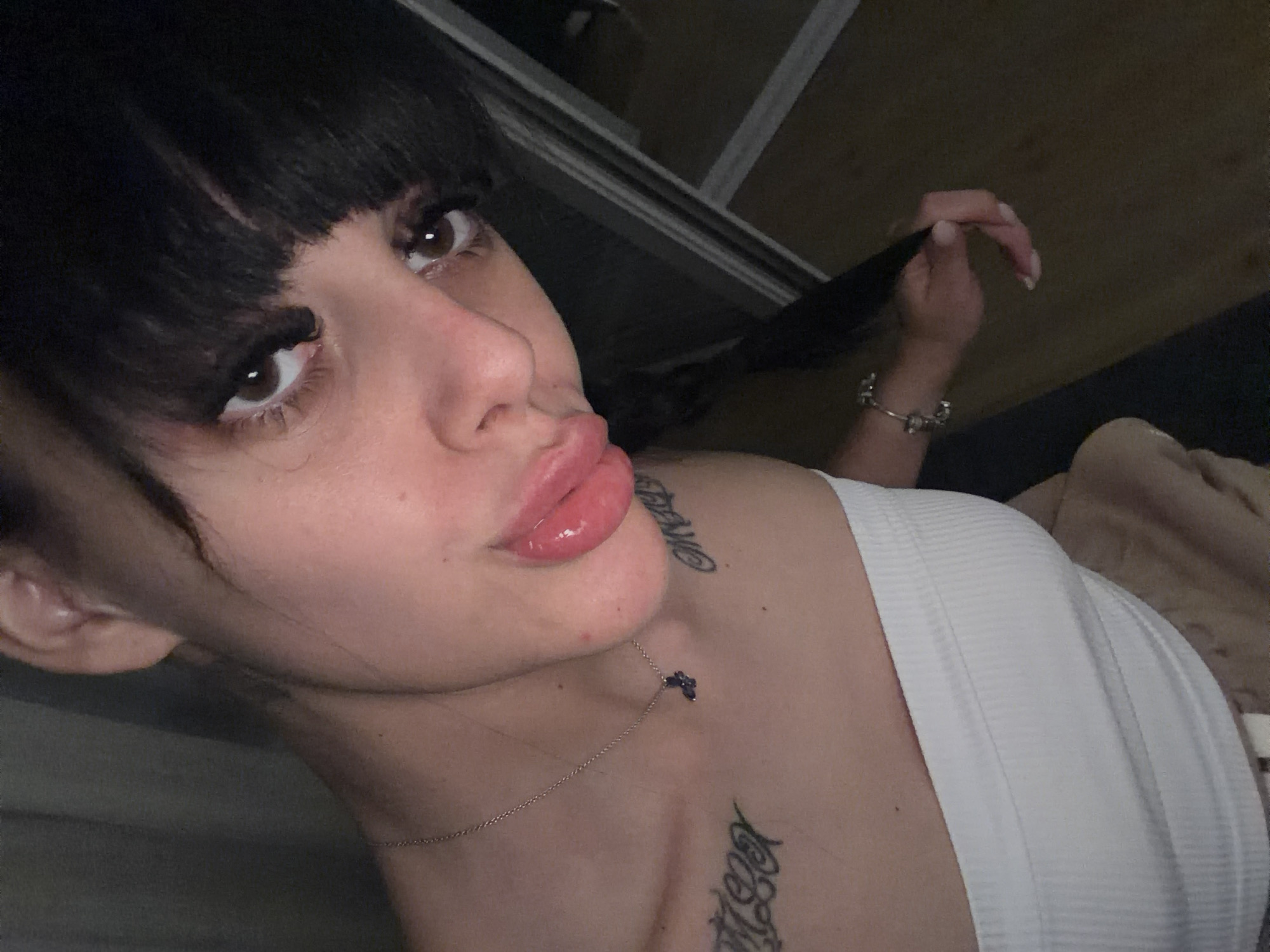 The image shows a close-up, high-angle selfie of a young woman with dark hair and bangs. She is wearing a white strapless top and a necklace with a small purple butterfly pendant. There are tattoos visible on her chest and collarbone. Her lips are slightly parted and painted with a glossy pink lipstick. Her eyes are brown and she is wearing false eyelashes. She is holding a strand of her hair in her right hand, which is adorned with a bracelet. The background is dimly lit and appears to be indoors, possibly a bedroom, with a dark curtain or wall on the left and a reflective surface or closet door on the right. The location in the city of Plovdiv, Bulgaria, is not discernible from the image.