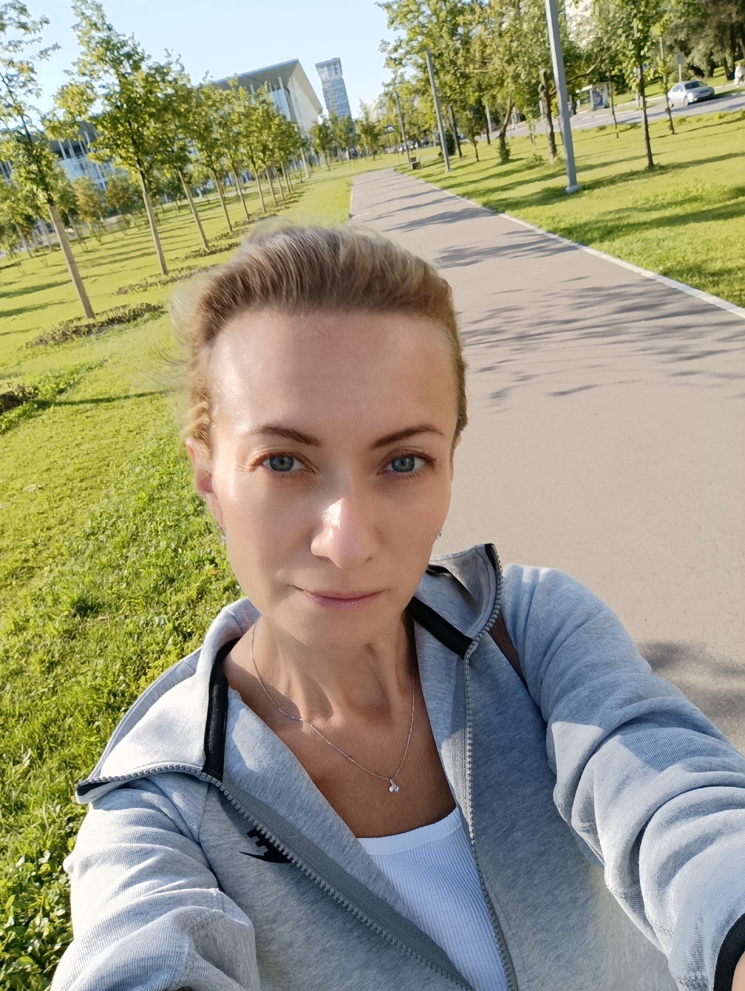 A medium shot selfie shows a woman with blonde hair pulled back, looking directly at the camera with blue eyes. She is wearing a grey zippered hoodie over a white ribbed tank top and a delicate silver necklace. The background depicts a paved path for walking or cycling, bordered by a well-maintained grassy area with young trees. In the distance, a modern building with glass facades and a tall tower are visible, suggesting an urban or suburban park setting in Moscow, Russia. The lighting indicates it is daytime, likely late afternoon, with shadows cast by the trees.