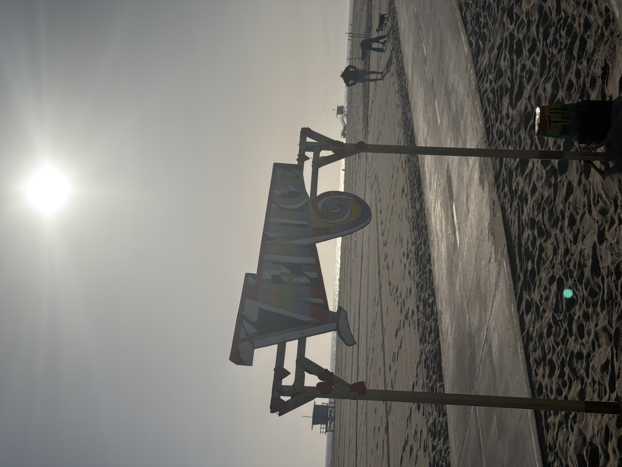 The photograph depicts a beach scene in Los Angeles, California, United States. The sun is high in the sky, creating a bright and hazy atmosphere. In the foreground, a colorful, geometrically shaped sign is mounted on wooden posts. In the background, the beach stretches into the distance, with a paved walkway adjacent to the sand. A few people can be seen walking or standing on the beach and the walkway. A lifeguard tower is visible in the distance. The beach is sandy, with visible footprints and tire tracks.