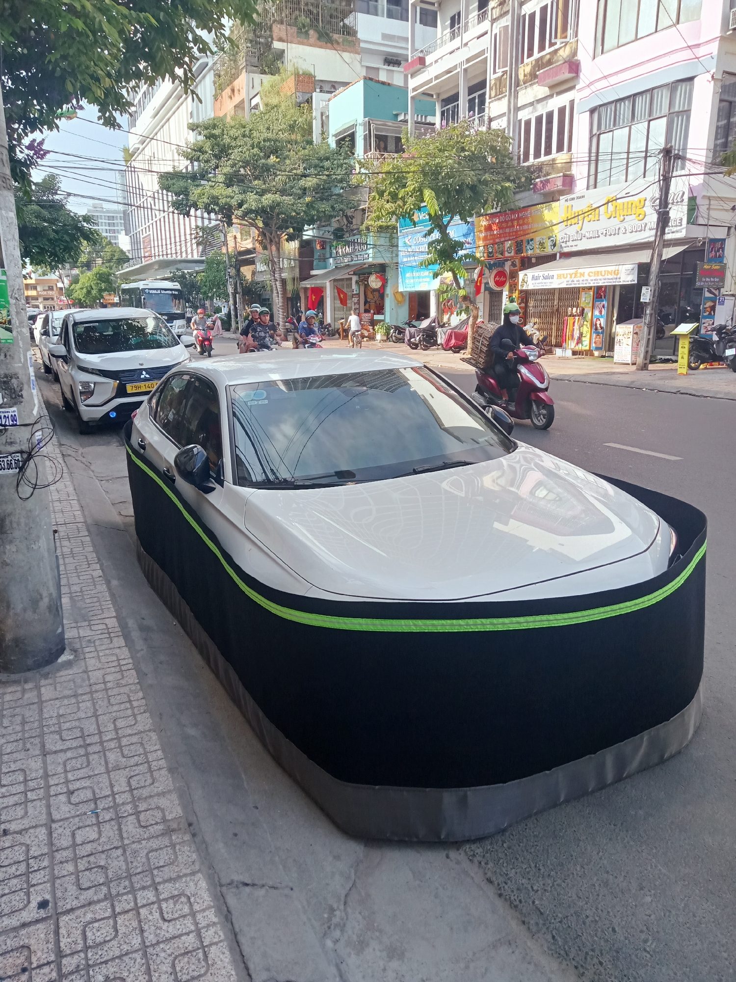 This daytime photograph captures a busy street scene in Nha Trang, Vietnam, featuring vehicles, people, and commercial buildings under a clear sky.
In the foreground, a white sedan is conspicuously parked on the side of the road, partially covered by a unique black fabric cover with a prominent lime green stripe around its middle. This cover extends to the ground, concealing the wheels and lower body of the car. Behind it, a white Mitsubishi Xpander with the license plate "79H-140" is also parked, followed by another white car. A light grey utility pole stands to the left.
The street is bustling with motorcycles, many carrying drivers and passengers. One person on a red scooter, wearing a face mask and carrying a large basket on the back, rides towards the right side of the frame. In the background, beyond the parked vehicles, a "VINAH Shuttle Bus" is partly visible.
The street is lined with multi-story buildings housing various businesses and residences, typical of an urban setting. Numerous trees provide shade along the sidewalk. Shop signs are visible, displaying Vietnamese text. Notable signs include "NÀNG ĐỒ GỖ MỸ NGHỆ N" (likely wood crafts), "HỘ KINH DOANH" (Business Household), and a yellow sign advertising "GỘI ĐẦU-NAIL-FOOT & BODY MICE" (Head wash-Nail-Foot & Body Massage). Further down, "Hair Salon HUYỆN CHUNG" is visible on a storefront. Other visible text fragments include "THÁNG LỢI", "21.99", "53.66.60", and an "Air Asia" logo on a smaller sign. The overall impression is one of a vibrant, active Southeast Asian city street.