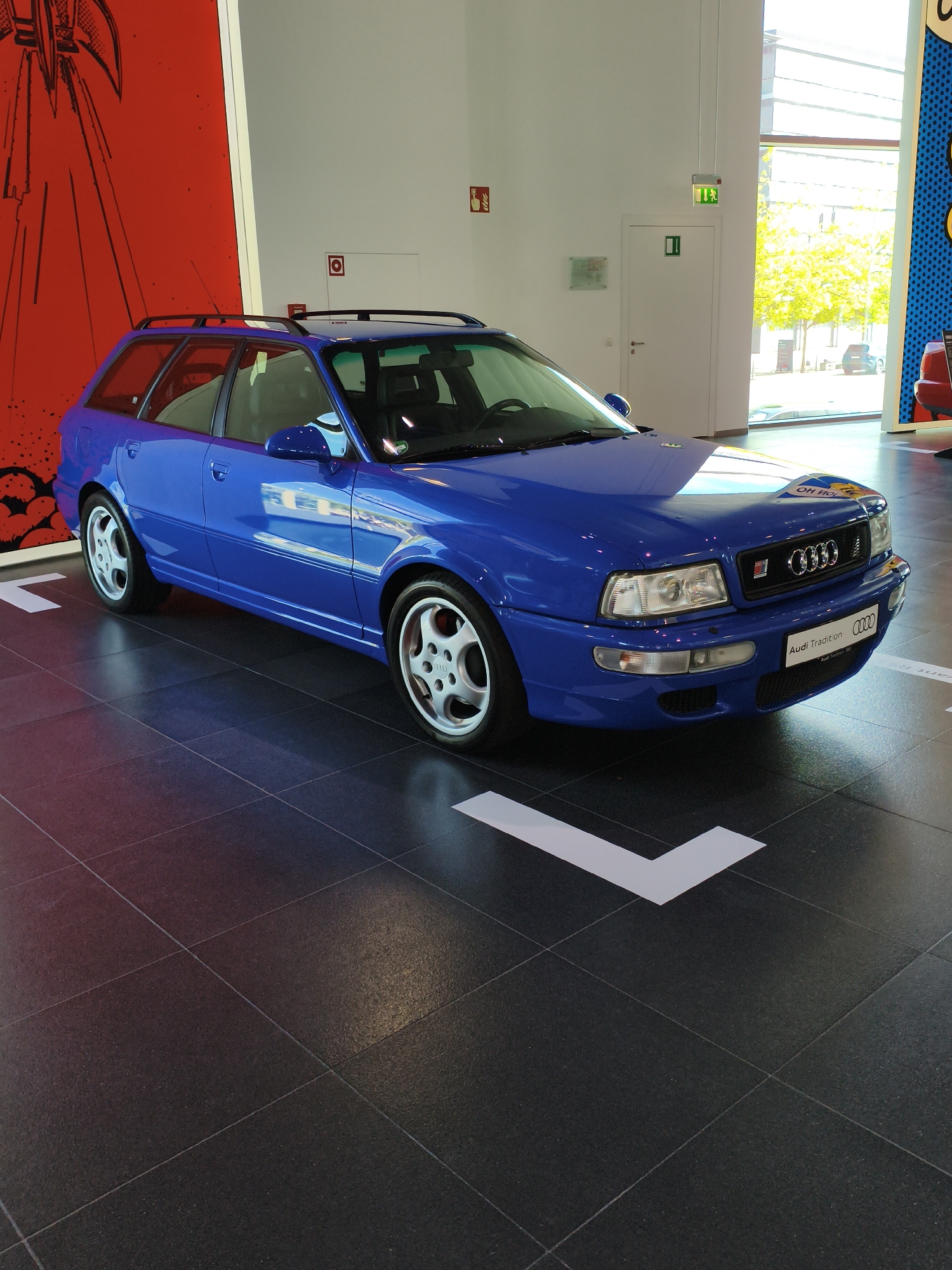 A bright blue Audi RS2 Avant is parked in a showroom or exhibition space. The car is positioned at an angle, facing towards the right of the frame. Its sleek design and vibrant color are highlighted by the lighting. The floor is made of large, dark grey tiles, some of which have white arrow-shaped markings. In the background, a large red abstract artwork with black lines is visible on the left wall. To the right, large windows allow natural light to enter, revealing trees and a cityscape outside. The overall environment appears to be a modern and well-lit space, possibly part of the Audi Museum Mobile in Ingolstadt, Germany, given the prominent display of the classic car. There are no people visible in the image, and the car is stationary.