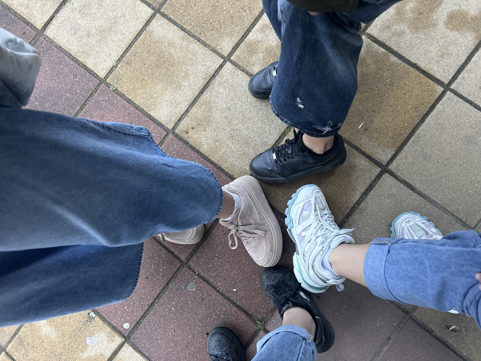 The image depicts a group of people standing on a tiled surface. The scene focuses on their lower legs and shoes. The individuals are wearing jeans of varying shades of blue. The shoes include a pair of black sneakers, light beige sneakers, a light-colored pair of Balenciaga sneakers and a pair of black water-covered sneakers. The tiled surface consists of squares of beige and red. The location of the photograph is unknown.