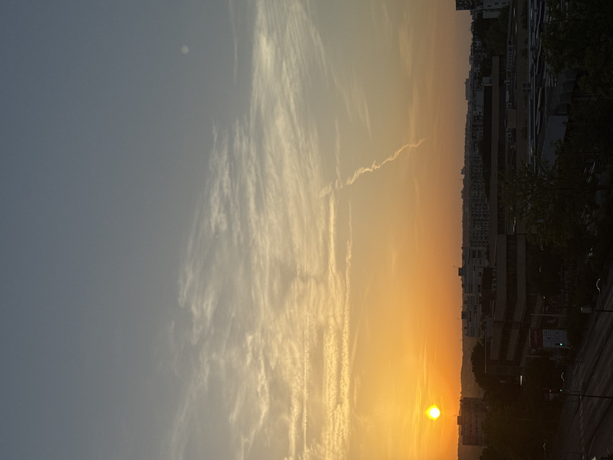 The image captures a sunset over a city skyline. The sky transitions from a pale blue on the left to a vibrant orange and yellow on the right, indicating the time of day is dusk. Wispy clouds streak across the sky, catching the warm light. A faint circular object, possibly the moon or a distant light source, is visible in the upper left portion of the sky.
In the foreground, silhouetted buildings and trees form the city's silhouette against the bright horizon. The architecture suggests a modern urban environment. A road with streetlights is visible on the right side of the image. The overall atmosphere is serene and peaceful, evoking the end of a day. There is no visible text in the image.