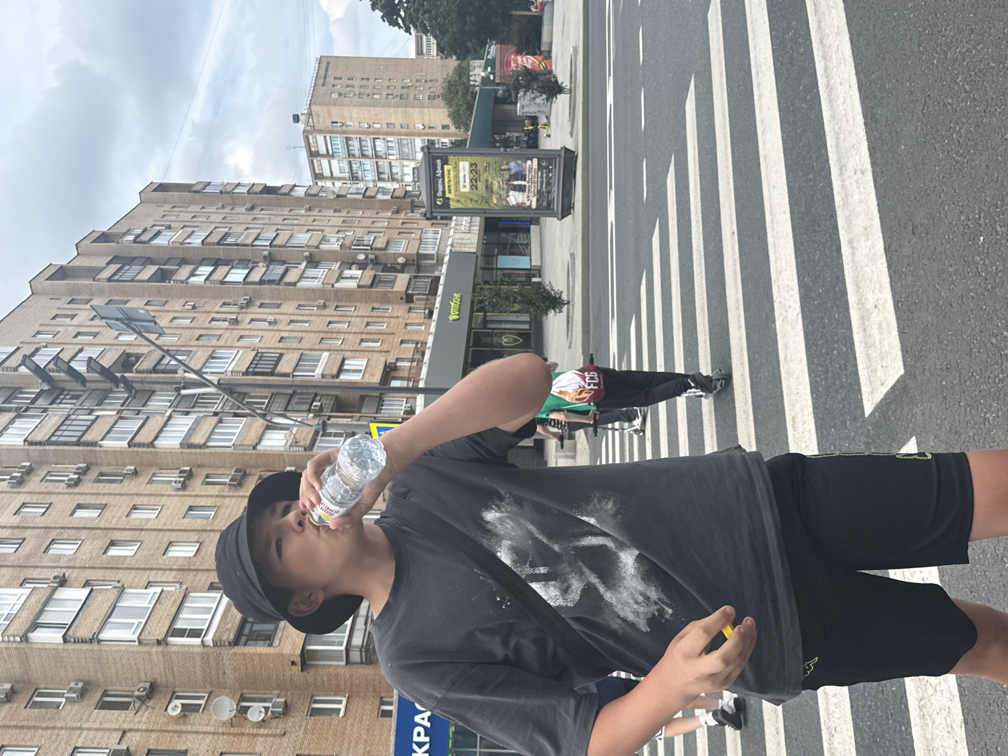 A young person wearing a black bucket hat and a dark t-shirt with a white graphic design is drinking from a clear plastic water bottle while standing on a pedestrian crosswalk in a city. They are wearing black shorts. In the background, there are tall brick apartment buildings with numerous windows and air conditioning units. To the right, a billboard advertising "Яндекс Афиша" is visible. Other people can be seen in the background, some with scooters. The sky is overcast. The scene appears to be in Moscow, Russia, given the "Яндекс Афиша" advertisement.
