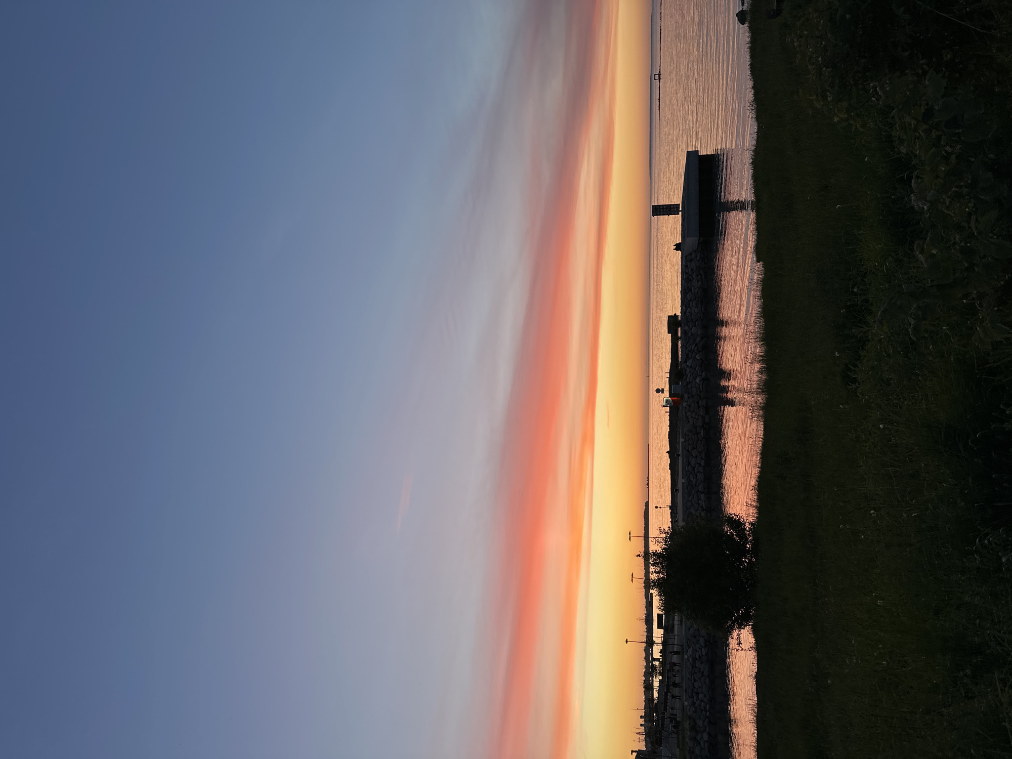 The image depicts a sunset over the Baltic Sea in Tallinn, Estonia. The sky is a gradient of pale blue at the top, transitioning to vibrant hues of orange and pink from the sunset. A thin strip of clouds stretches across the horizon, illuminated by the setting sun. In the foreground, a grassy area is silhouetted against the twilight. Beyond the grass, a body of water reflects the colorful sky. A stone pier extends into the water, with a few indistinct figures visible on it. Further in the distance, the skyline of Tallinn is partially visible, with some buildings and cranes silhouetted against the sky. The overall impression is one of a peaceful evening by the sea.