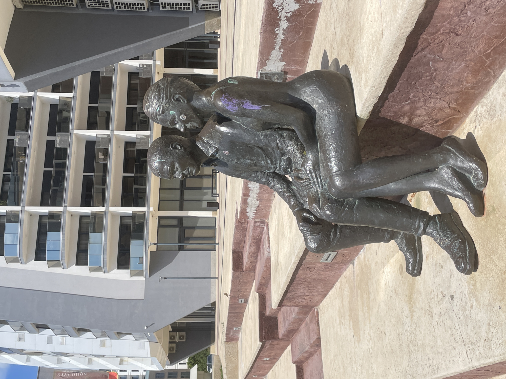 The image shows a bronze sculpture depicting two men in an embrace, located in Bar, Montenegro. The sculpture is positioned on a stepped, light-colored concrete base with reddish-brown accents. A modern, multi-story building with glass windows and gray and white facade is visible in the background. The sculpture is slightly marred by purple paint on the back of one of the figures.