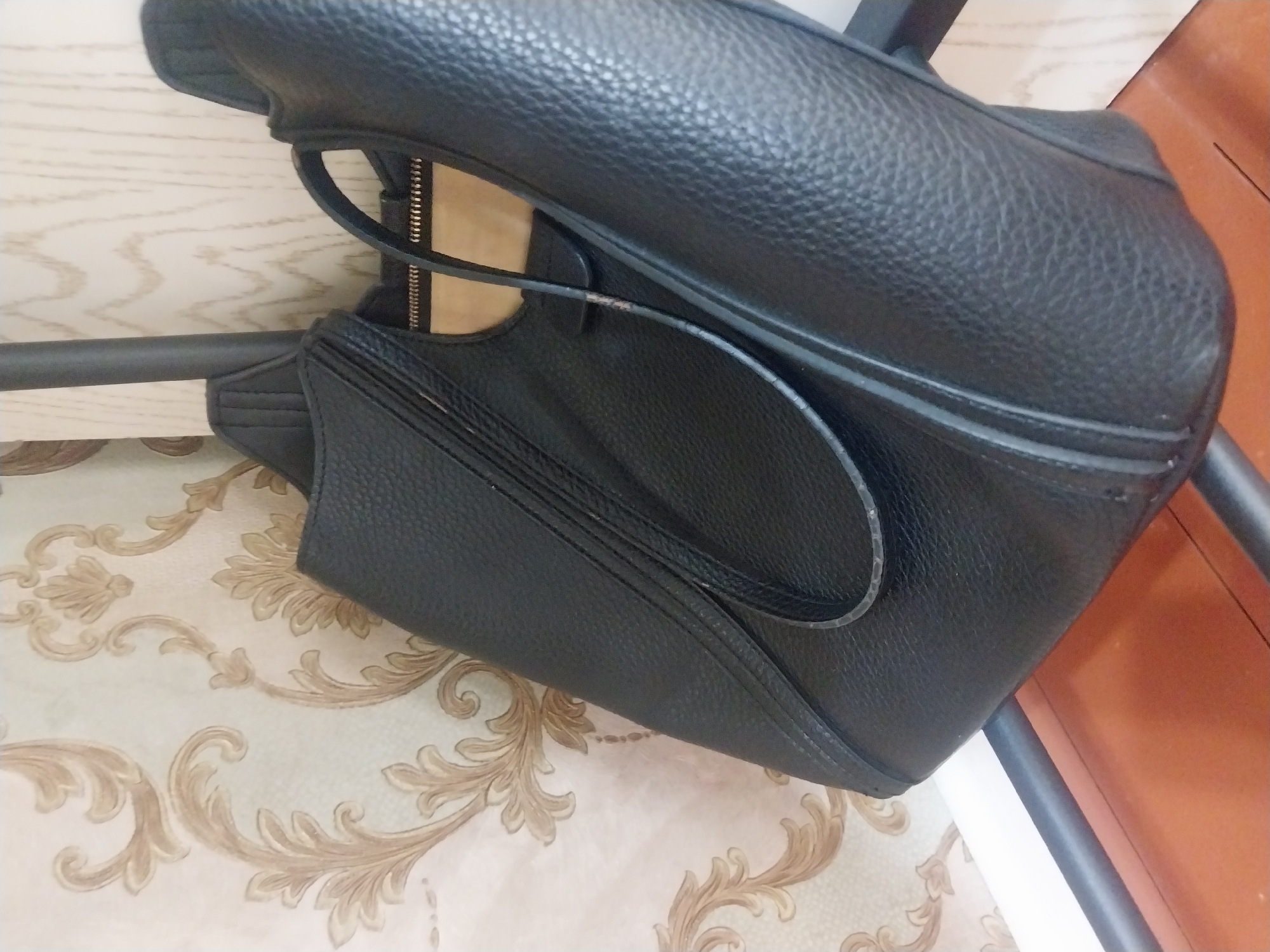 A black, textured leather tote bag hangs on a dark metal pole, angled towards the right side of the frame. The bag is partially open, revealing a beige interior and a silver zipper. The background consists of a light-colored wallpaper with a gold, ornate floral pattern, and a section of light wood grain. The bag is positioned in front of a white baseboard. Part of a wooden surface is visible at the bottom of the image. The scene appears to be indoors, possibly in a domestic setting. The city of Blagoveshenka, Kyrgyzstan, is not depicted in this image.