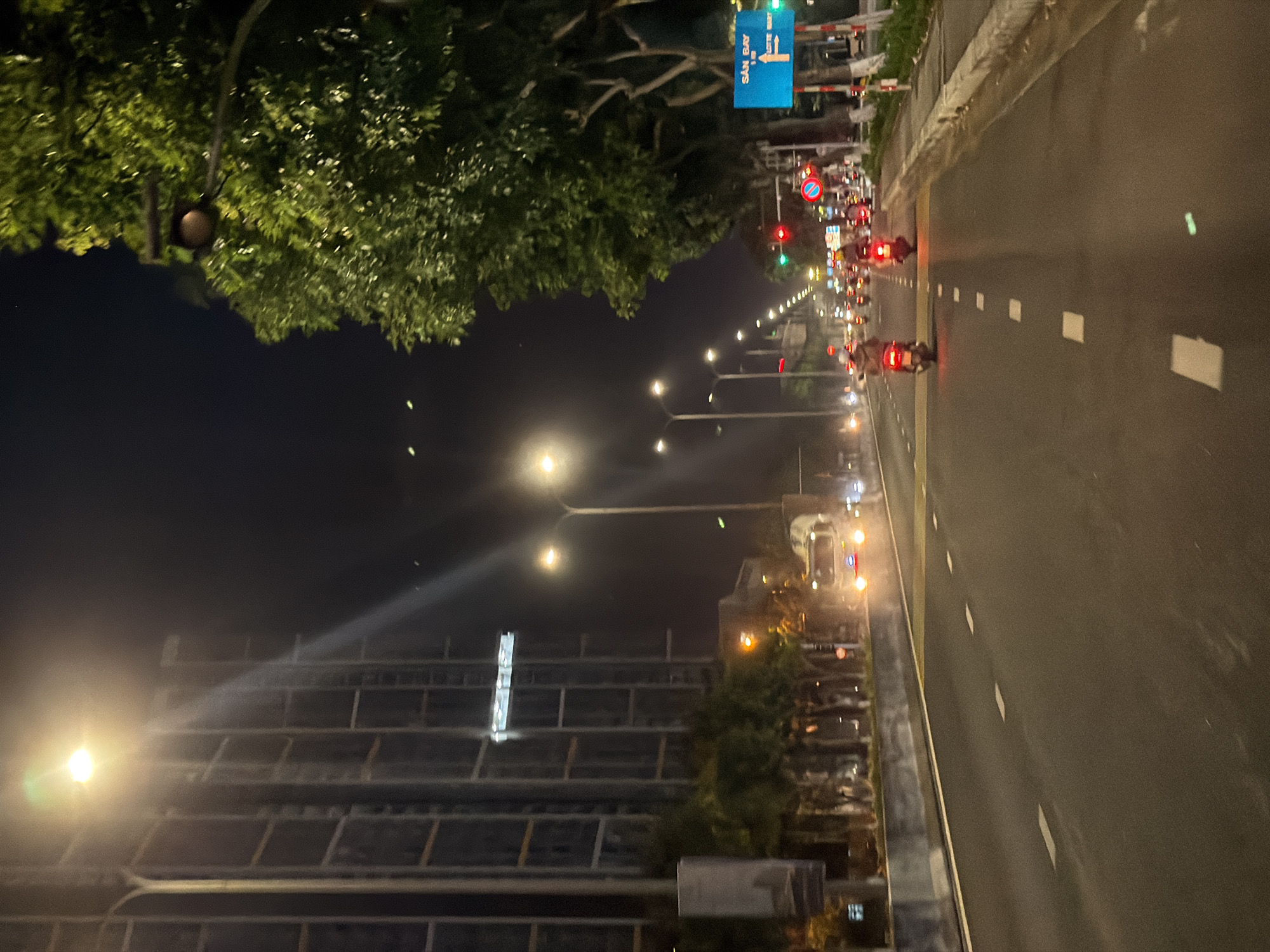 The image shows a street scene at night in Da Nang, Vietnam, likely on a road with multiple lanes. A tall, modern building with a grid-like facade is visible on the left, illuminated by streetlights. To the right, lush green trees line the street, with a street lamp visible through the branches.
The road itself is dark asphalt with white dashed lane markers. Several motorcycles are present, some with riders. One motorcycle in the foreground has its taillight illuminated. Further down the road, a truck with its headlights on is visible. Traffic lights are showing red and green signals. A blue street sign with white text and an arrow indicating directions is also visible on the right side of the road. The overall atmosphere is urban and dynamic, typical of a city at night with ongoing traffic.