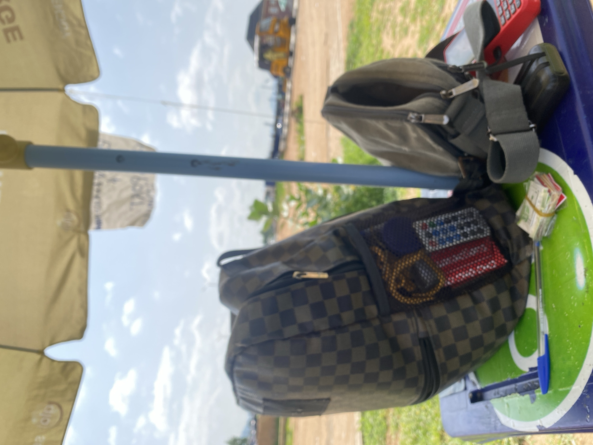 The image depicts an outdoor scene, possibly a street market stall in Jalingo, Nigeria. In the foreground, two backpacks are placed on a blue and green table. The larger backpack, on the left, has a checkered pattern in shades of brown and green. A mesh pouch on this backpack contains a keychain with a gold-colored fob and a small item resembling a wallet or pouch decorated with red, white, and blue patterns. To the right of the larger backpack is a smaller, plain gray backpack with a strap.
On the table, a blue pen lies on the green surface. To the right of the gray backpack, a stack of banknotes, secured with yellow rubber bands, is visible. Further to the right, a red calculator is partially obscured by the gray backpack. A black object, possibly a phone, is positioned beneath the gray backpack strap.
In the background, a yellow three-wheeled vehicle, commonly known as a "Keke Napep" in Nigeria, is driving on a dusty road. The environment appears to be an open area with dry vegetation and a few buildings in the distance. The sky is partly cloudy. An umbrella, likely yellow, is casting a shadow over the scene, suggesting it is daytime. There are no people clearly visible in the image.