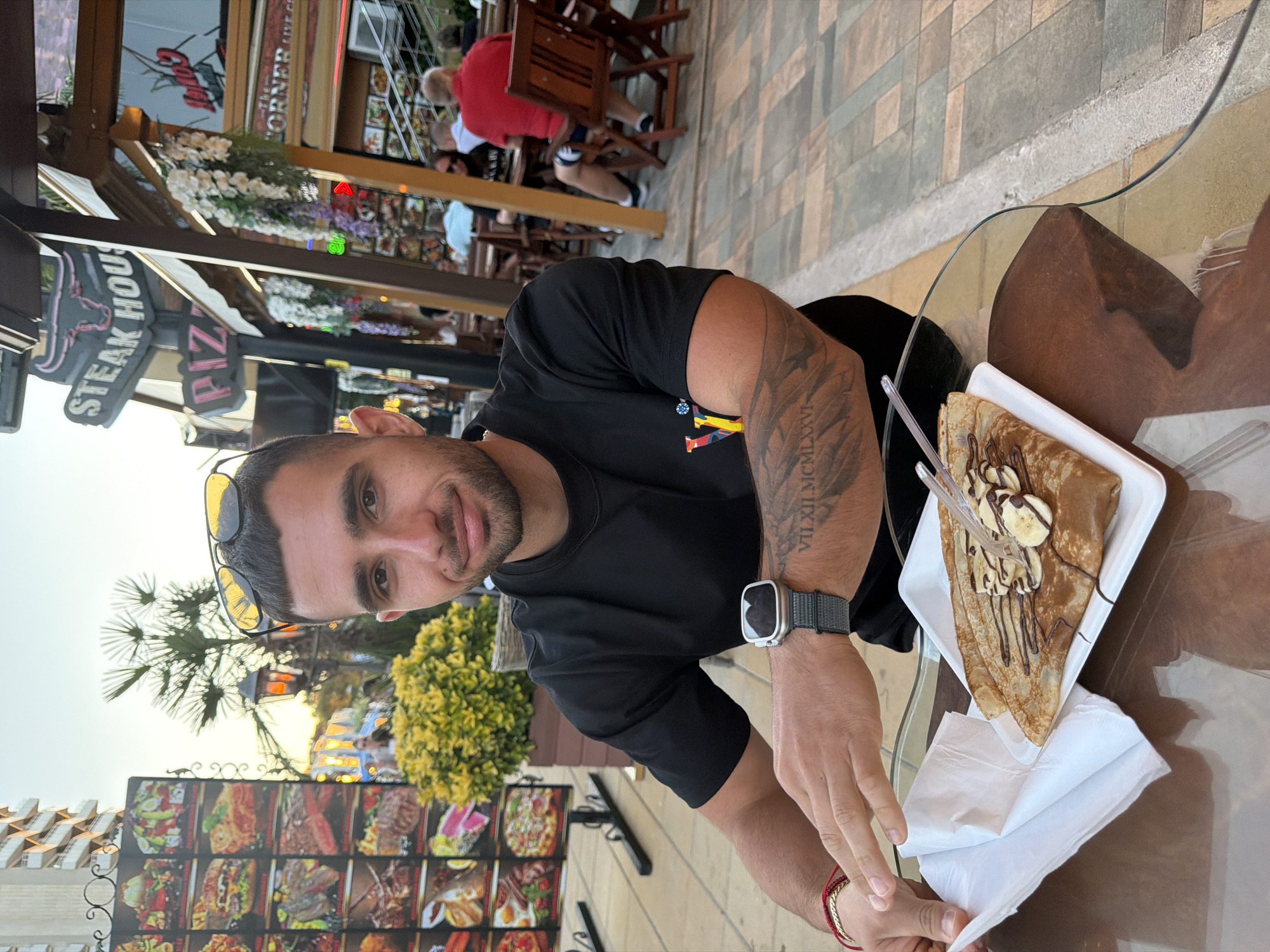 A man with short dark hair and a beard is seated at a table outdoors, looking towards the camera. He is wearing a black t-shirt and a watch on his left wrist. In front of him is a crepe with chocolate drizzle and banana slices on a white plate, with a plastic fork resting on it. A napkin is to the left of the plate. The table has a glass top and a wooden base. In the background, there are other tables and people dining, as well as restaurant signage that reads "STEAK HOUSE" and "PIZZA". A building with balconies is visible in the far background. The man has tattoos on his left arm and a bracelet on his left wrist. The scene appears to be in an outdoor dining area in Nesebar, Bulgaria, with palm trees and decorative plants visible. The lighting suggests it is late afternoon or early evening.