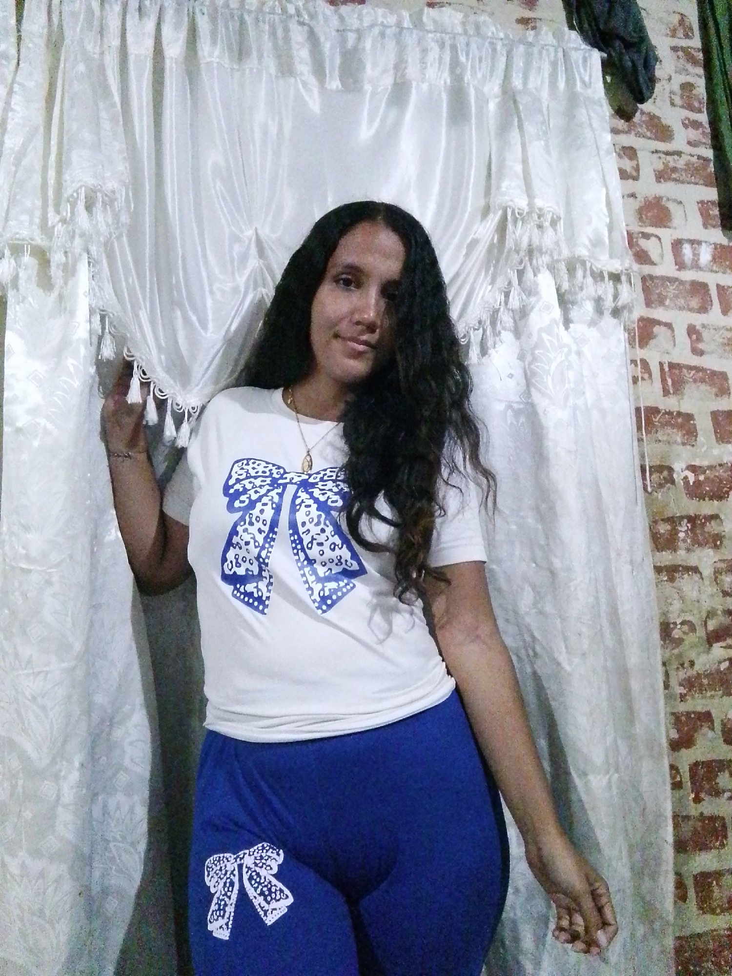 A woman stands indoors, posing for a photograph. She has long, dark, curly hair and wears a white t-shirt with a large blue bow print on the front and blue leggings, also with a bow print on the leg. The background includes a light-colored curtain behind her and a brick wall to her right. The specific location is stated as Juan Griego, Venezuela.