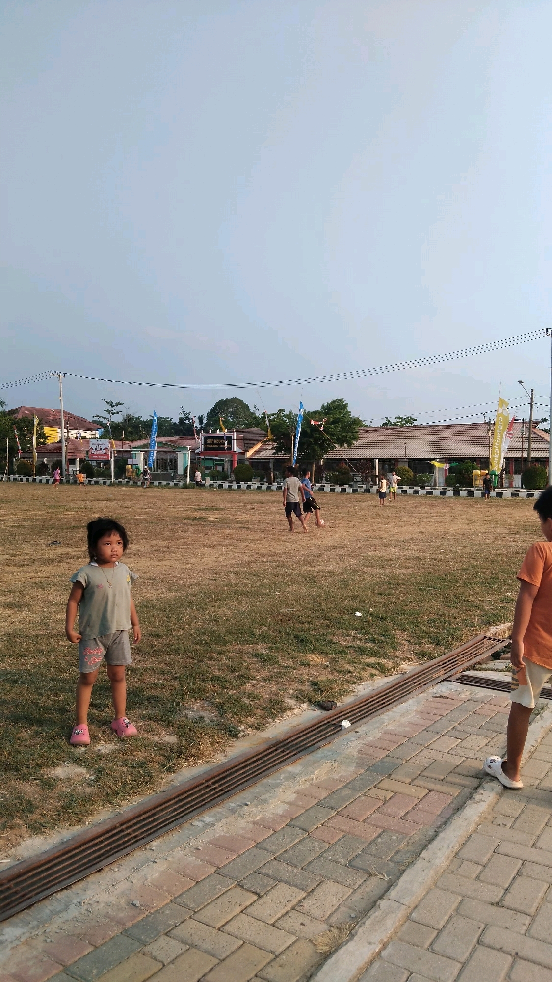 In Manunggal Jaya, Indonesia, a dry, grassy field is the setting for an informal soccer game among several boys. In the foreground, a young girl, around three years old, stands facing left, looking slightly upwards with a curious expression. She wears a light grey t-shirt with a graphic on it and grey shorts, complemented by pink sandals. To the right, a boy wearing an orange t-shirt and white shorts stands facing the game, his back mostly to the camera.
Behind the children, a school building with a tiled roof and painted walls is visible, adorned with various flags and banners. The field is bordered by a black and white striped fence. Power lines and poles are present in the background, and the sky is a pale, overcast blue. The ground along the edge of the field features a brick pathway and a metal drainage grate. The overall scene depicts a casual outdoor activity in a community space.