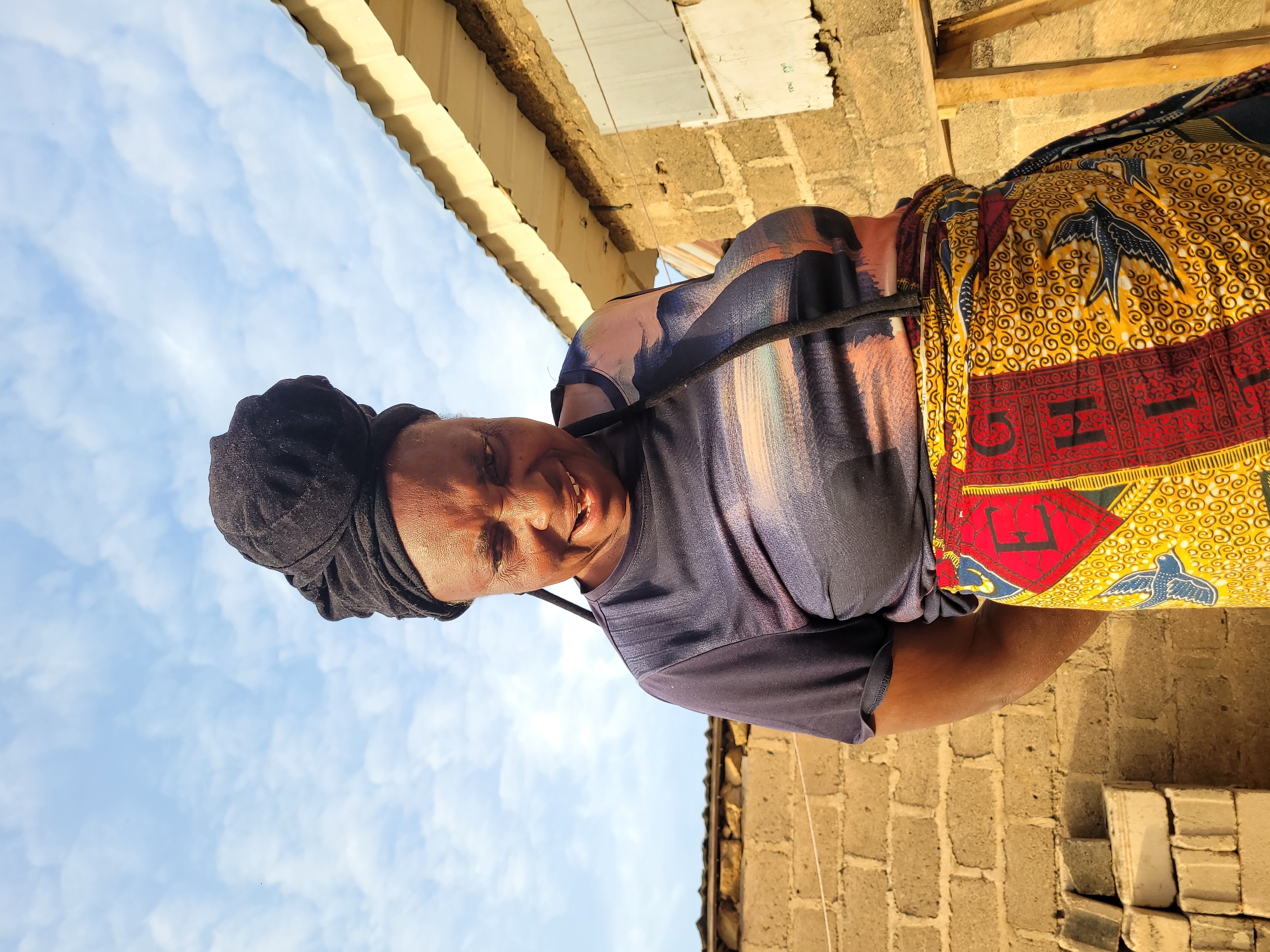 The image captures a dark-skinned woman smiling directly at the camera, set against a bright blue sky with scattered white clouds. She is positioned slightly to the left of the frame, and the photograph is taken from a low angle, emphasizing the sky and the top portion of a nearby building.
The woman wears a black headwrap. Her top is a short-sleeved, blue or dark grey garment with subtle abstract patterns in lighter shades. A dark strap or rope crosses her chest. She is dressed in a vibrant, full skirt or wrap that features a yellow background with intricate black swirling patterns and stylized black birds resembling swallows. The bottom section of the skirt has a prominent red band adorned with repeating patterns and includes distinct black letters, notably "G" and "H," along with an "X O" border design.
In the background, elements of a building are visible, constructed from concrete blocks. A corrugated iron roof and wooden support beams are seen above the woman. To her right, more of the concrete block wall is present, and at the bottom right, a stack of white bricks or building blocks lies on the ground. The clear sky and bright illumination suggest it is daytime, likely sunny. The woman's expression is one of warmth and friendliness. While some faint markings are visible on parts of the building in the upper background, they are not clearly legible. The scene's construction style and natural light cues are consistent with the provided location context of Barakin Akawo, Nigeria.