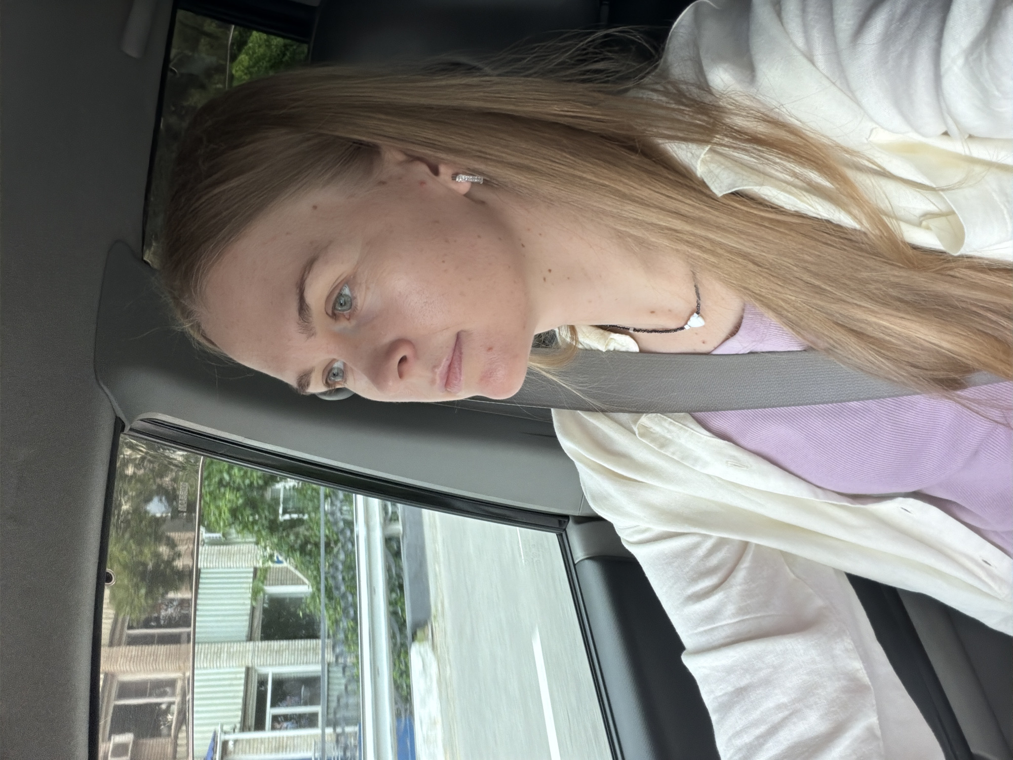 A blonde woman with blue eyes is driving a car on a street in Khabarovsk, Russia. She is wearing a light purple t-shirt and an open white button-up shirt. She is looking to her left, out the window. Buildings and trees are visible outside the car window. The seatbelt is visible across her chest. She has on a necklace and earrings.