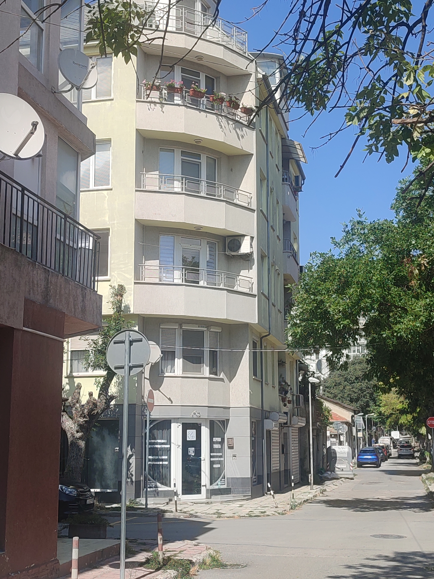 This is an outdoor, daytime, eye-level, medium shot of a street in Varna, Bulgaria. The scene is dominated by apartment buildings and trees. A multi-story apartment building with balconies and satellite dishes is on the left. Another similar building is on the right, with a slightly curved facade. Several cars are parked on the street, and one is driving away from the camera. The street is paved with asphalt, and there are sidewalks on both sides. The sky is clear and blue. The image appears to have been taken on a sunny day.