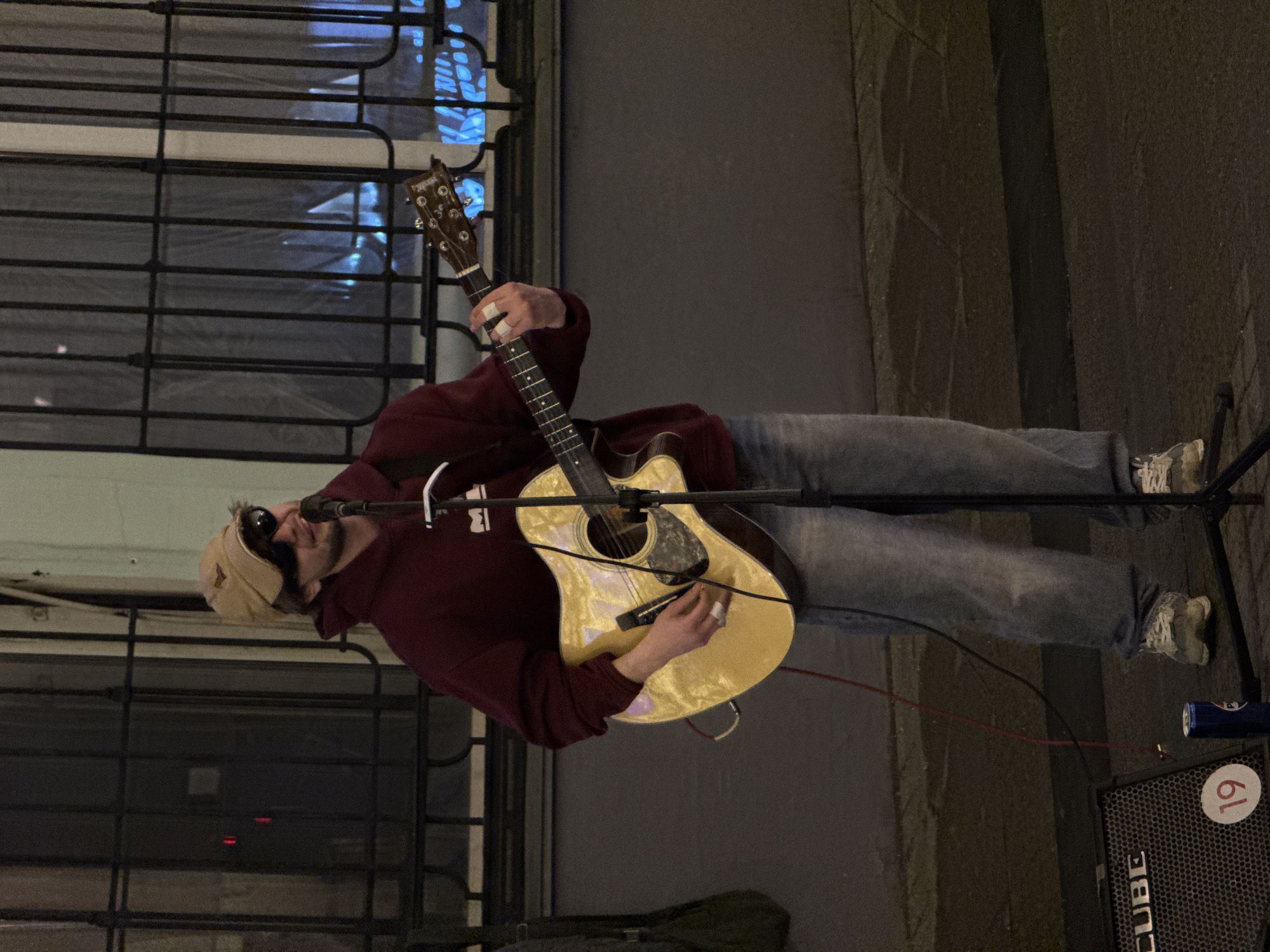 The image depicts a street musician performing outdoors in an urban environment. The musician is a man wearing a dark red hoodie, jeans, and sneakers, and a tan cap. He is playing an acoustic guitar and singing into a microphone that is mounted on a stand. He appears to be wearing sunglasses. In front of him is a guitar amplifier labeled "Cube". There is an object with the number "19" on the top right of the amp. The setting appears to be alongside a building with windows that have a grid-like pattern. Based on the window style and the overall architectural feel, it is plausible that the location is within Moscow, Russia, though this is speculative without further evidence. The ground is paved.