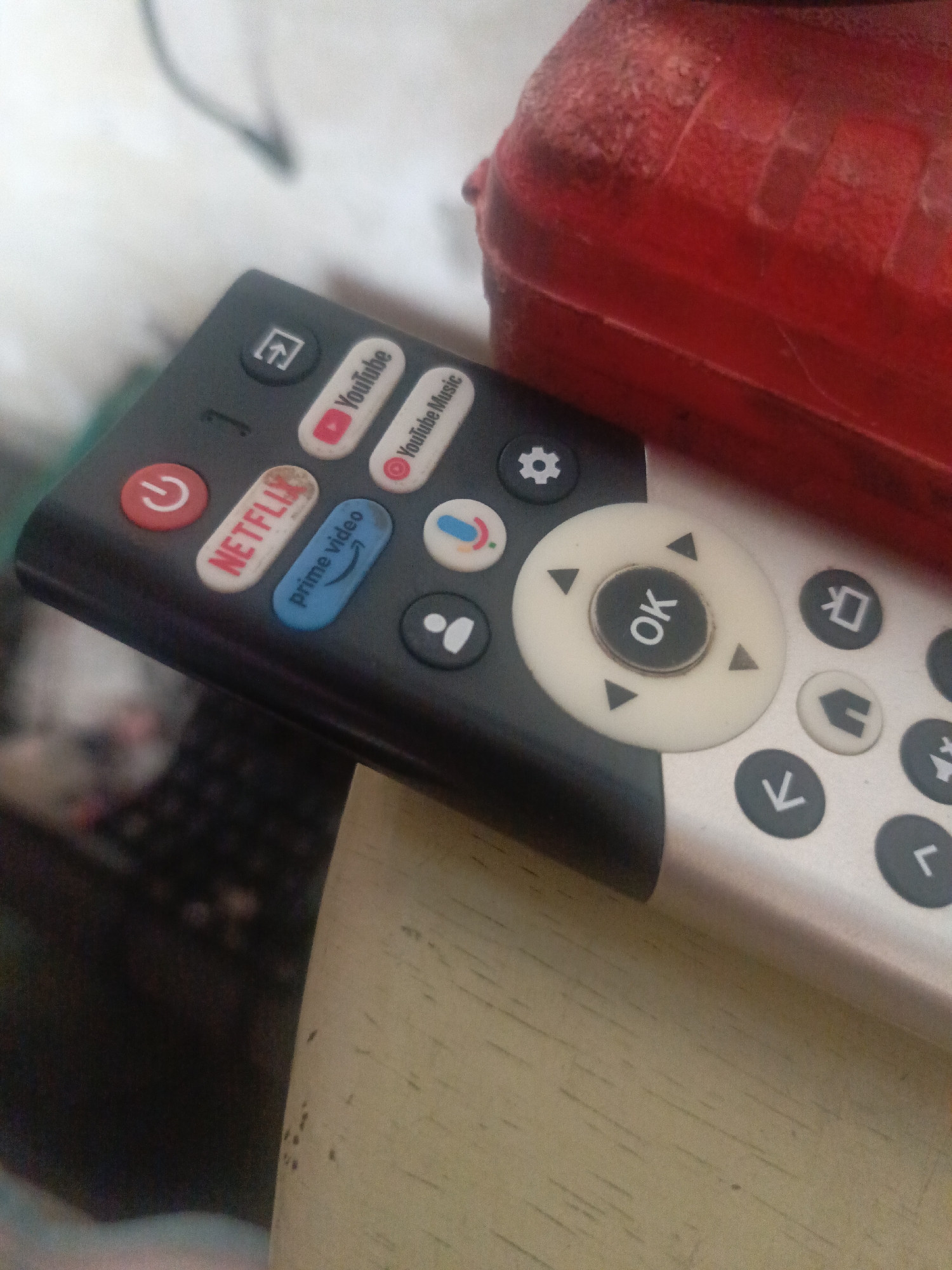 A close-up shot shows a black and silver remote control with buttons for Netflix, YouTube, Prime Video, and other streaming services. The remote is resting on a surface with light tan color. A red object is partially visible in the upper right corner of the frame. The image does not depict any discernible location in the city of Juan Griego, Venezuela, nor does it include details about people, actions, or interactions.