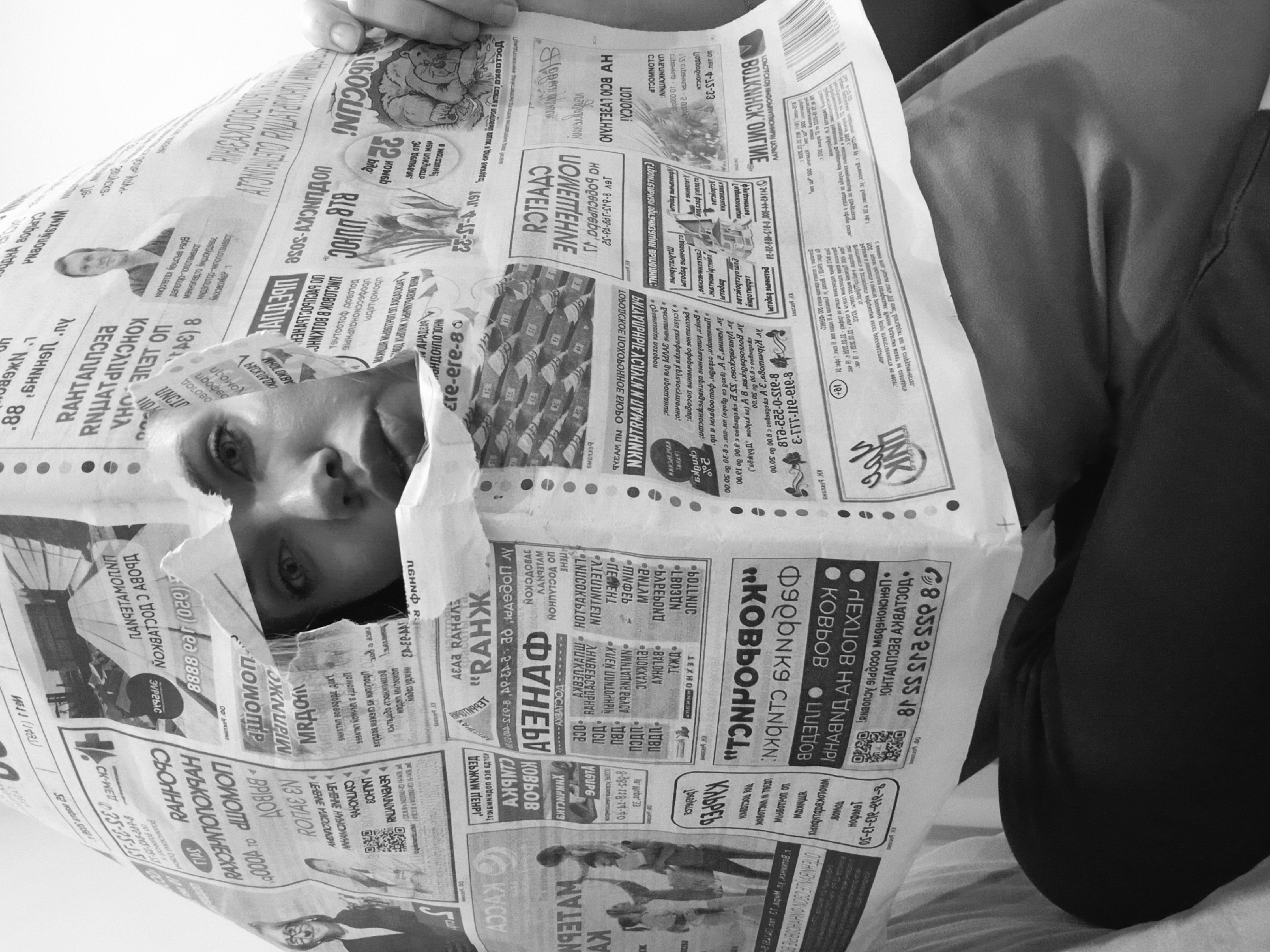 A grayscale medium shot depicts a person with a newspaper held up to their face. The newspaper is mostly in Cyrillic and obscures much of the person’s face except for their eyes, nose, and part of their mouth. The newspaper is held in front of them by a hand at the top. The person is wearing a dark garment. The newspaper’s text includes the name "Votkinsk" in Cyrillic.