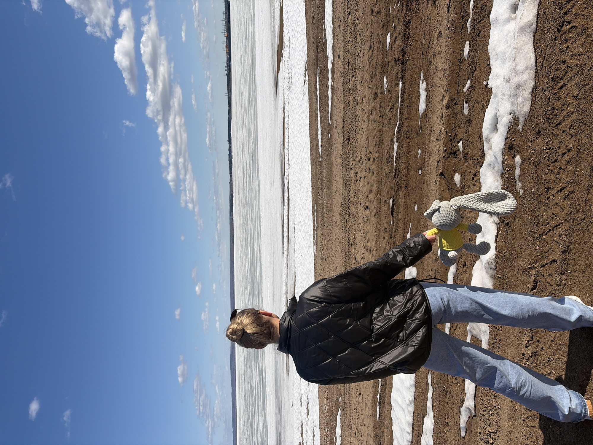 The image shows a person standing on a beach near the town of Noviy, Russia. The person is facing away from the camera, towards a frozen body of water. The sky is blue with some clouds. The beach is covered in dirt with streaks of snow. The person is wearing a black jacket and light blue jeans. They are holding a stuffed rabbit toy.