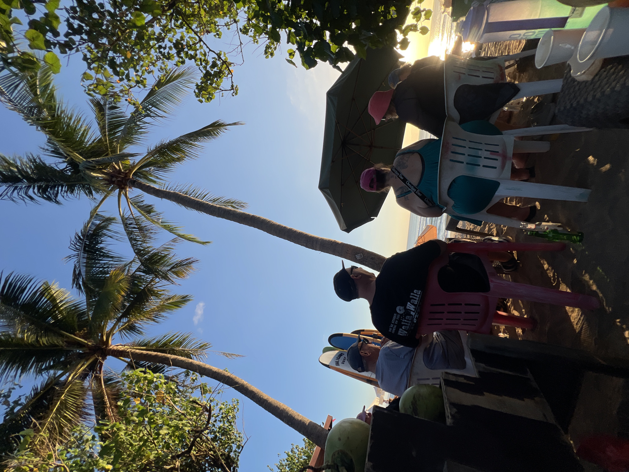 The image depicts a beach scene in Kuta, Indonesia, with two tall palm trees framing a clear blue sky. Several people are seated on plastic chairs facing the ocean, which is visible in the background with the sun low on the horizon. In the foreground, there is a wooden structure with coconuts on it, and surfboards leaning against it. An umbrella is set up over some of the seated individuals. The ground is sandy, with some green bottles and cups visible on the beach. The overall atmosphere suggests a relaxed, end-of-day setting.