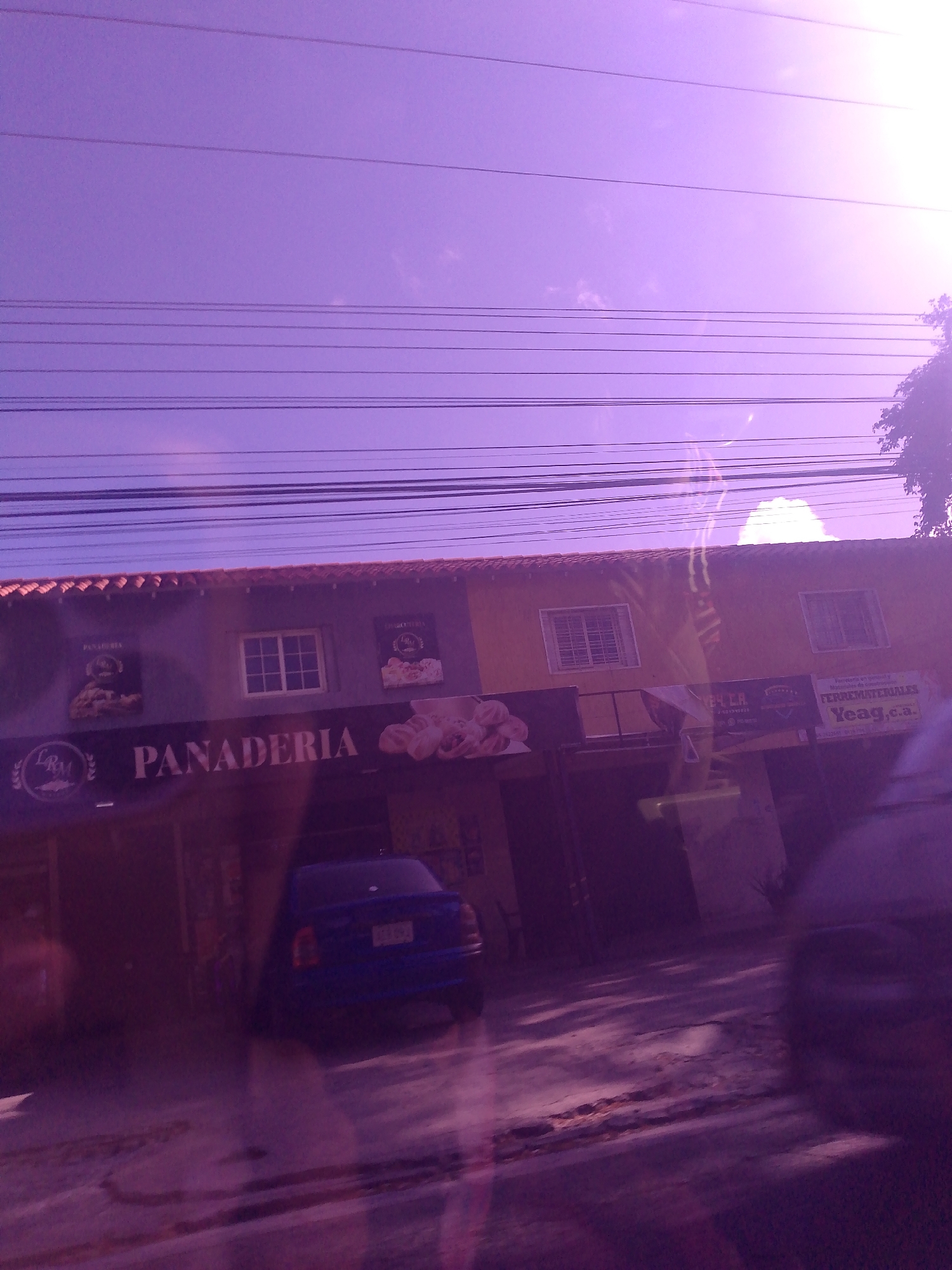 The image depicts the front facade of a one-story building in La Asuncion, Venezuela, identified by a sign reading "PANADERIA." A blue car is parked in front of the bakery. Overhead, numerous electrical or telephone wires are visible against the bright sky. A small tree is seen on the right. The image appears to have been taken through a window or windshield, as evidenced by reflections and potential water streaks on the glass.