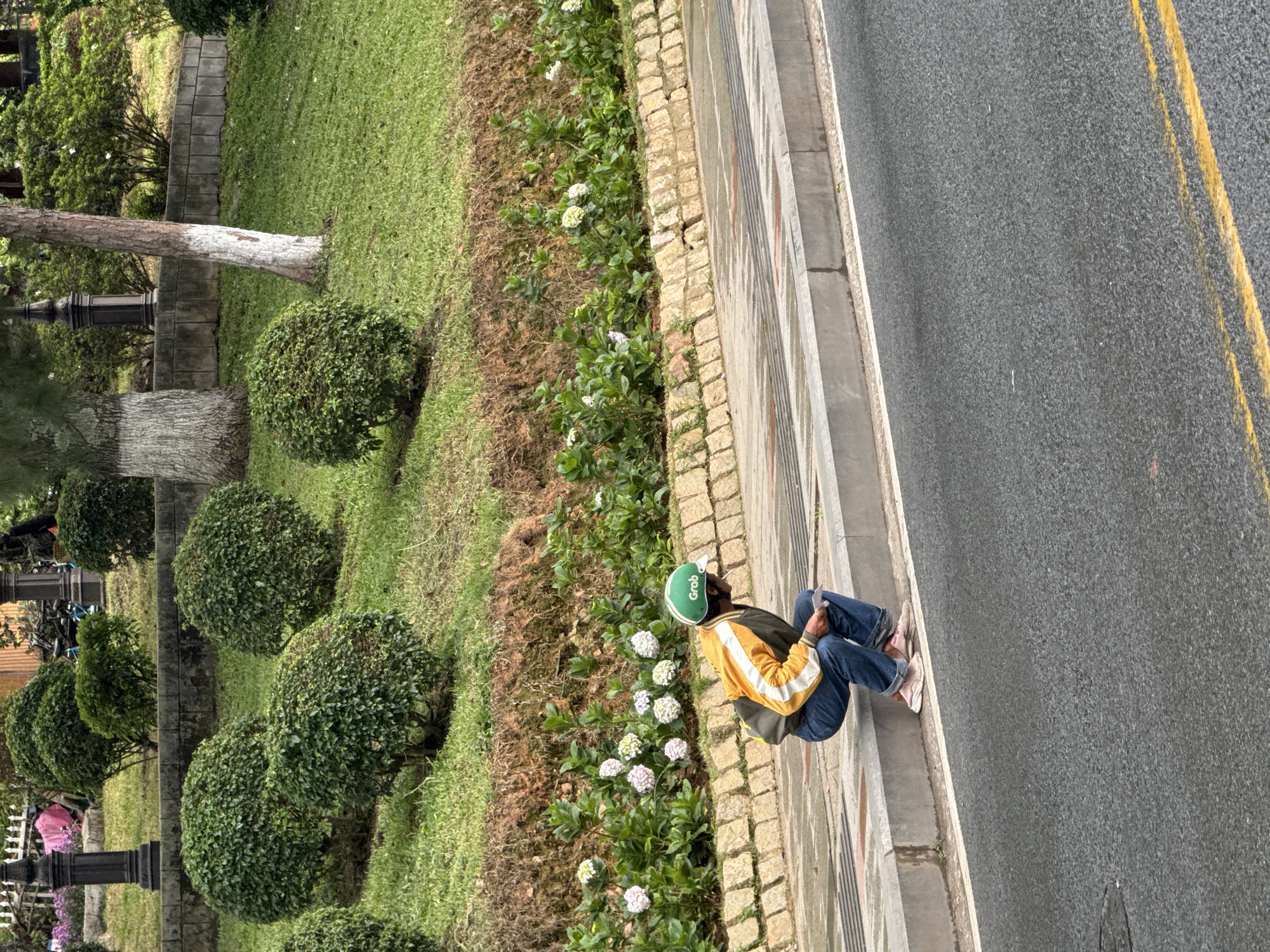 A person wearing a green helmet with "Grab" printed on it, a yellow and white jacket, and blue jeans squats by the side of a road in Da Lat, Vietnam. The person is holding a piece of paper. The road is asphalt with yellow lines. A concrete curb separates the road from a grassy area with manicured bushes and flowering plants. In the background, a stone wall and more vegetation are visible.