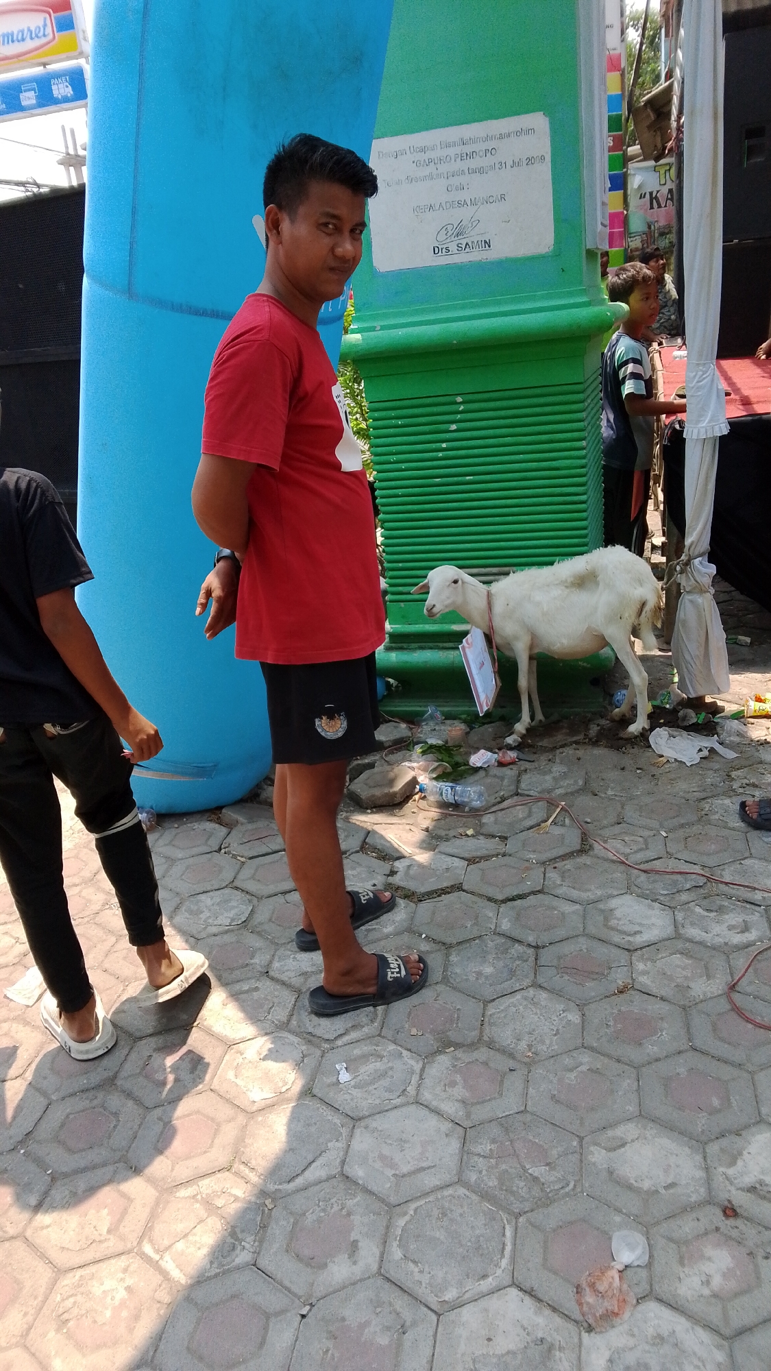 The image depicts an outdoor scene on a paved hexagonal tile surface, likely in Mancar, Indonesia, judging by the inscription on a green structure. A young man wearing a red t-shirt and black shorts stands near a large, inflated blue structure. To his right, a white goat is tethered and has a sign hanging around its neck. In the background, a green structure with horizontal ribbing is visible, bearing text on a white plaque. This plaque reads, "Dengan Ucapan Bismillahirohmannirohim 'GAPURO PENDAPO' Telah diresmikan pada tanggal 31 Juli 2009 Oleh: KEPALA DESA MANCAR Drs. SAMIN". Several plastic bottles and wrappers are scattered on the ground around the goat and the green structure, indicating a possibly recent event or ongoing activity. Another young person in a black t-shirt and black pants is partially visible to the left of the man in the red shirt, facing away from the camera. Other individuals and structures are present in the background, suggesting a public gathering or event. The overall lighting suggests it is daytime.