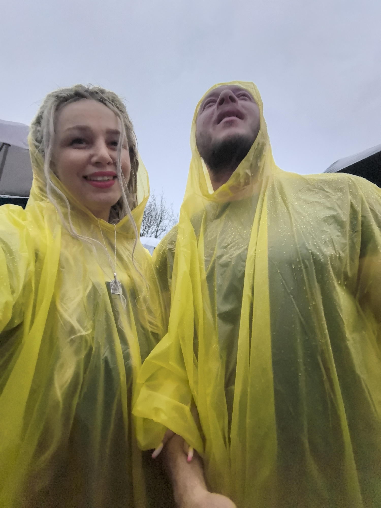 The image shows a close-up, waist-up view of a couple dressed in bright yellow rain ponchos, standing outdoors on an overcast day. The woman on the left is smiling and looking towards the camera. She has blonde dreadlocks and is wearing a silver necklace with a rectangular pendant. The man on the right is looking upwards with his mouth slightly open, as if reacting to something above. Both are wearing dark clothing underneath their ponchos, which are speckled with raindrops. The background is blurred and suggests a slightly wooded or natural area with bare trees visible. The overall atmosphere is casual and appears to be a candid shot taken during rainy weather. There is no visible indication of the specific location within Nizhny Novgorod, Russia.