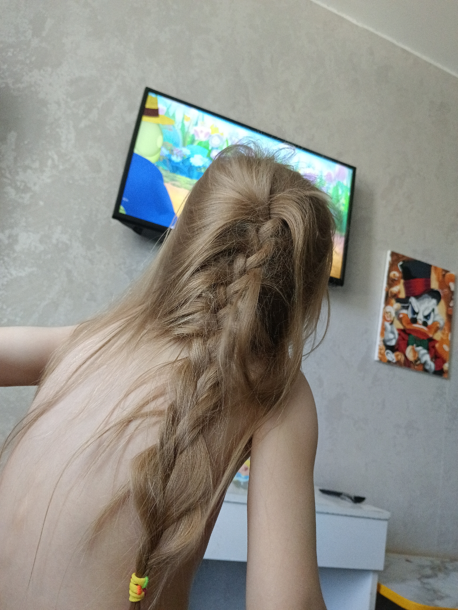A person with light brown hair, styled in a braid, is facing away from the camera. The braid is secured at the end with a small, colorful hair tie. The person is indoors in a location in the city of Chaikovsky, Russia. A television screen showing a cartoon is mounted on the wall behind them. To the right of the television, there is a picture featuring a cartoon duck character. Below the television and picture, there is a white piece of furniture with a black object on top. The walls are light gray.