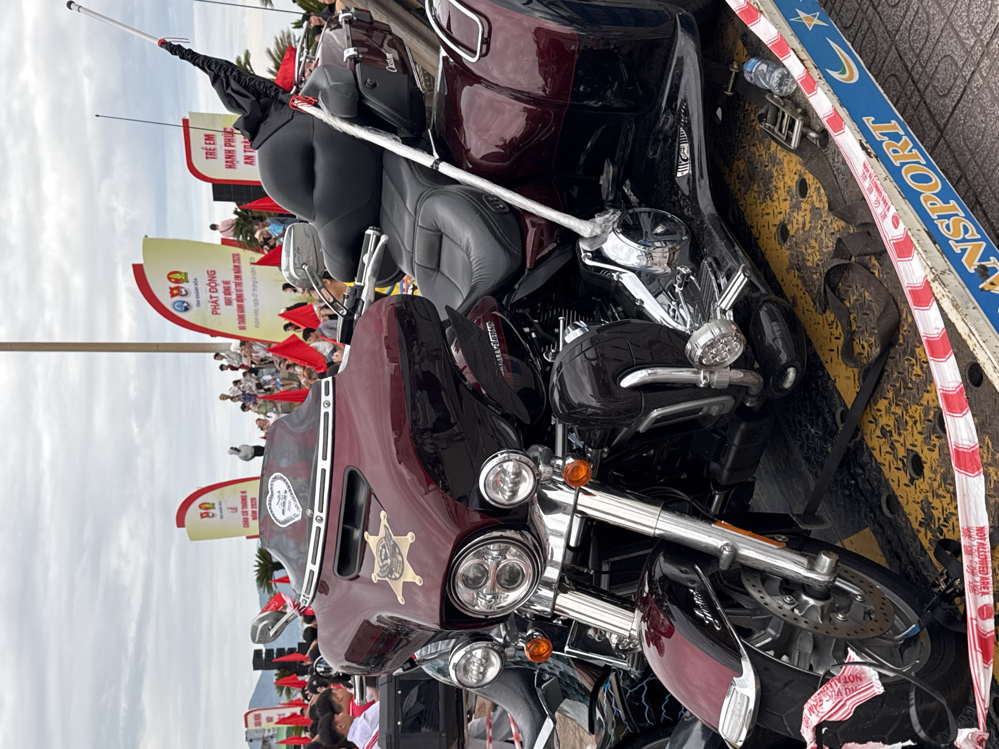 The image depicts a maroon and black Harley-Davidson motorcycle secured on the back of a flatbed truck. The bike features a sheriff star emblem on its fairing. A blue and white "ANSPORT" sign is visible along the truck's edge. A crowd of people, some holding small red flags, stands in the background near two signs in Vietnamese. The scene is likely in Nha Trang Ward, Vietnam, based on the signage. The sky is overcast. Other motorcycles are visible near the primary subject.