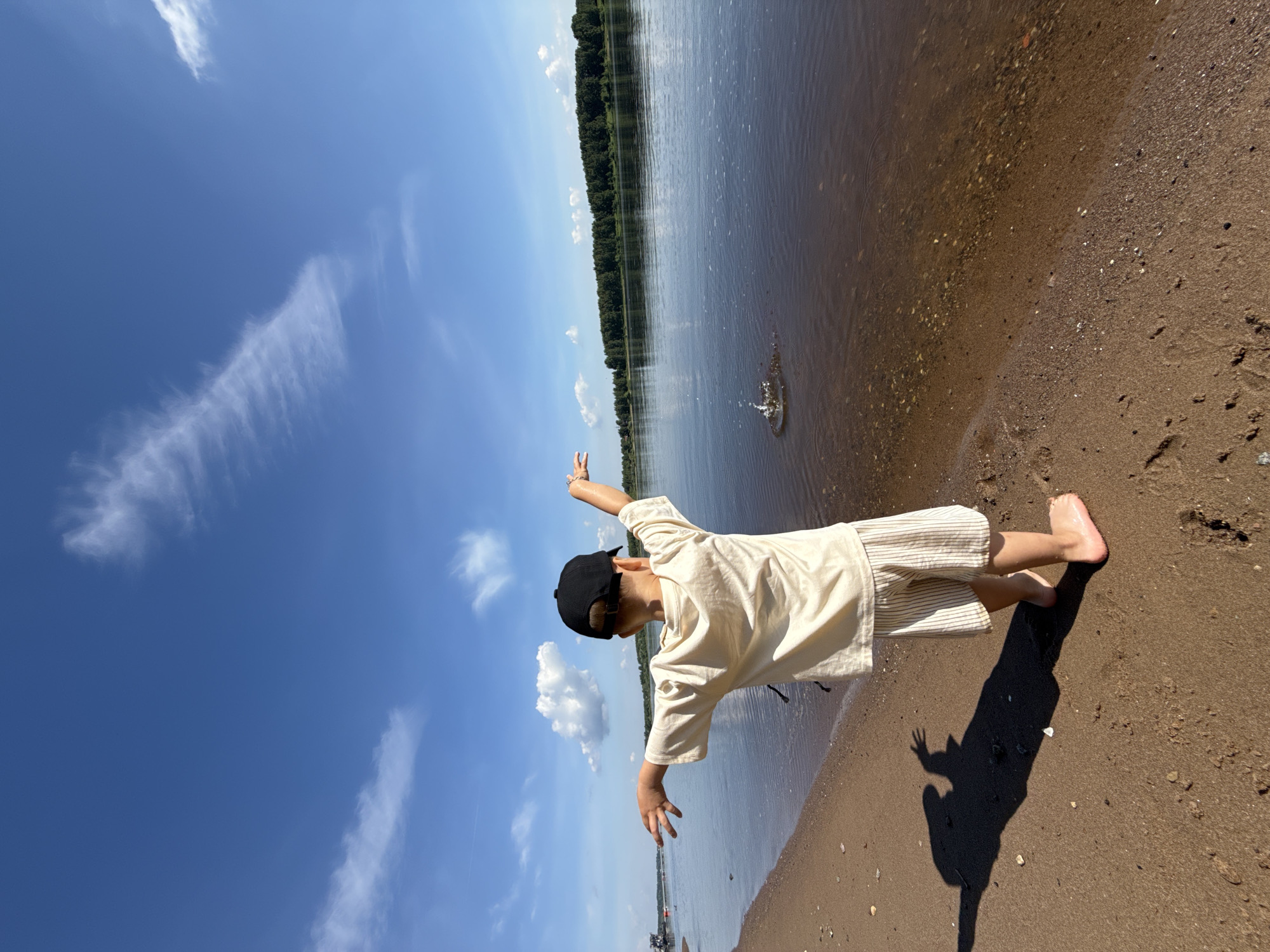 A young child wearing a light-colored t-shirt and striped shorts, with a black baseball cap, stands on a sandy shore with their arms outstretched towards a calm body of water. The child's shadow is cast on the sand. A splash is visible in the water, indicating an object has been thrown. In the background, a tree-lined horizon stretches across the landscape, with a blue sky filled with scattered white clouds above. The location is the bank of the Volga River in the city of Zavolzhye, Russia.