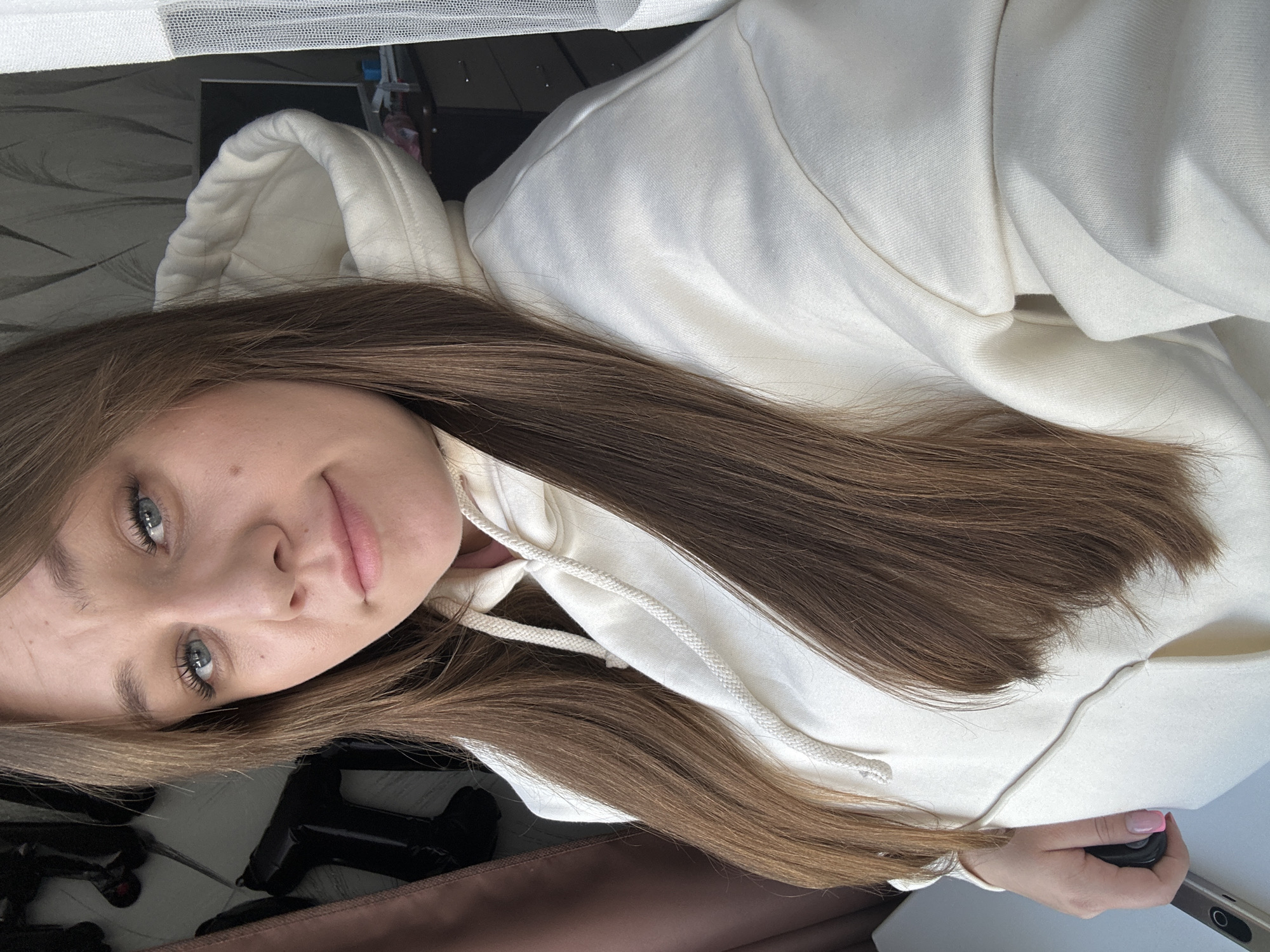The scene is an indoor residential setting located in Chaikovsky, Russia. A young adult female is depicted in a close-up, chest-up portrait. She possesses fair skin, light blue eyes, and long, straight brown hair that extends over her shoulders. The subject is wearing a cream-colored hooded sweatshirt featuring a visible drawstring. Her left hand, with fingernails painted pink, is visible in the lower left portion of the frame, grasping a dark-colored electronic device. She maintains direct eye contact with the camera, exhibiting a subtle, closed-mouth expression.
The background on the left side shows a brown curtain or drape. Partially obscured by the subject's hair, several black, irregularly shaped objects, possibly inflatable decorations, are affixed to a light-colored wall with a subtle pattern. On the right side of the frame, a sheer white curtain is visible. Behind the subject's right shoulder, a dark-toned dresser or cabinet with multiple drawers is present, topped by a dark flat-screen monitor and other small, indistinct items. The wall behind the dresser features a patterned wallpaper, possibly gray with light, vertical designs. The lighting in the room is ambient, consistent with an indoor environment.