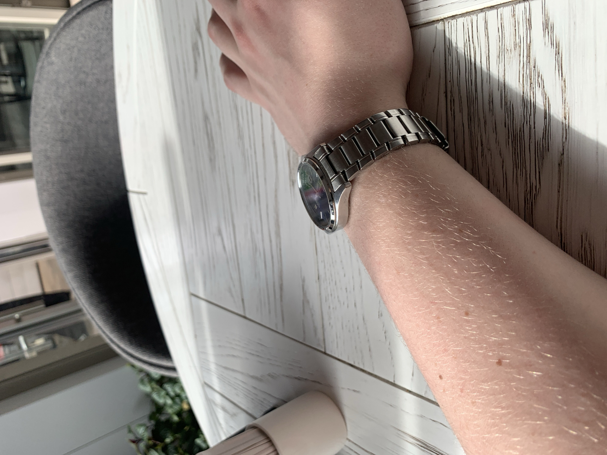 The image depicts a person's arm with a silver wristwatch resting on a white wooden surface. The arm is positioned on a table, and a gray chair is visible in the background. The background also includes glimpses of outdoor greenery, suggesting a location near a window or an outdoor area. There is no information about the city of Almaty, Kazakhstan, so this cannot be determined from the image.