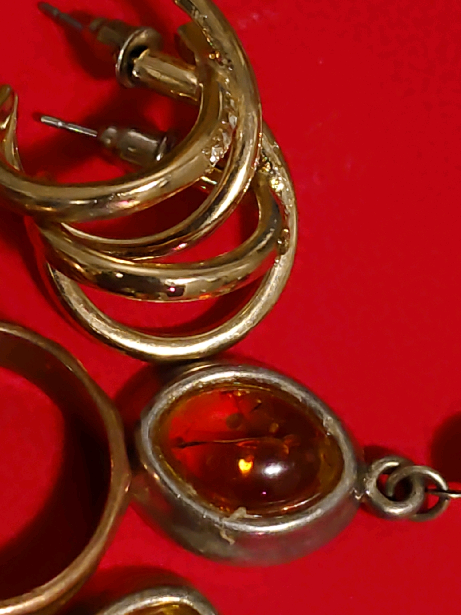 This is a close-up, overhead shot of several gold-colored jewelry pieces against a bright red background. The jewelry includes hoop earrings with visible posts and clutches, a ring with a large, oval, amber-colored stone set in a silver-colored metal band, and another, smaller ring or charm. The lighting creates reflections on the polished surfaces of the gold and the facets of the amber stone. The red background appears to be a fabric, possibly velvet, with some slight texture visible. There are no people or actions depicted in the image. The location is not discernible from the image itself.