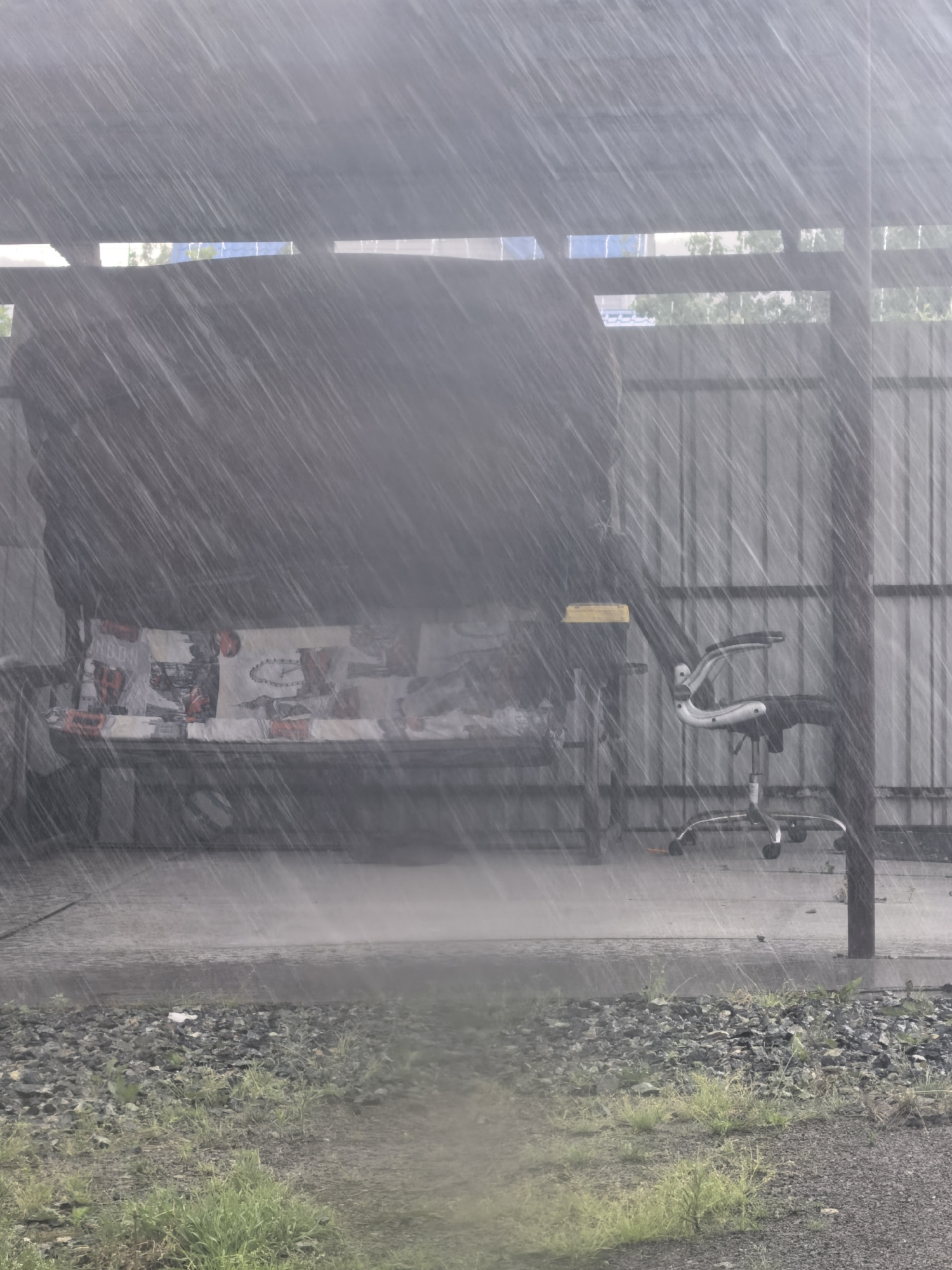 This media file captures an outdoor scene during heavy rainfall. The primary subjects are a couch with a patterned fabric and an office chair with a metal base. The couch is positioned under a covered structure, suggesting a sheltered outdoor area, possibly a patio or carport. The office chair is located to the right of the couch, also under the cover.
The scene is dominated by the weather event: the image is filled with streaks of rain, creating a blurred and hazy effect. The overall atmosphere is one of a downpour, with water visibly splashing on the ground in front of the couch. The ground itself is a mix of gravel and sparse grass. Behind the covered area, a corrugated metal fence is visible, providing a backdrop to the scene.
The lighting suggests it is daytime, but the heavy rain obscures details and creates a muted, gray palette. There are no people visible in the image, and the focus is entirely on the objects and the weather. The setting appears to be utilitarian or perhaps slightly neglected, given the presence of outdoor furniture and a metal fence. The strong visual of the rain creates a sense of a natural event in progress. No text is visible in the media file.