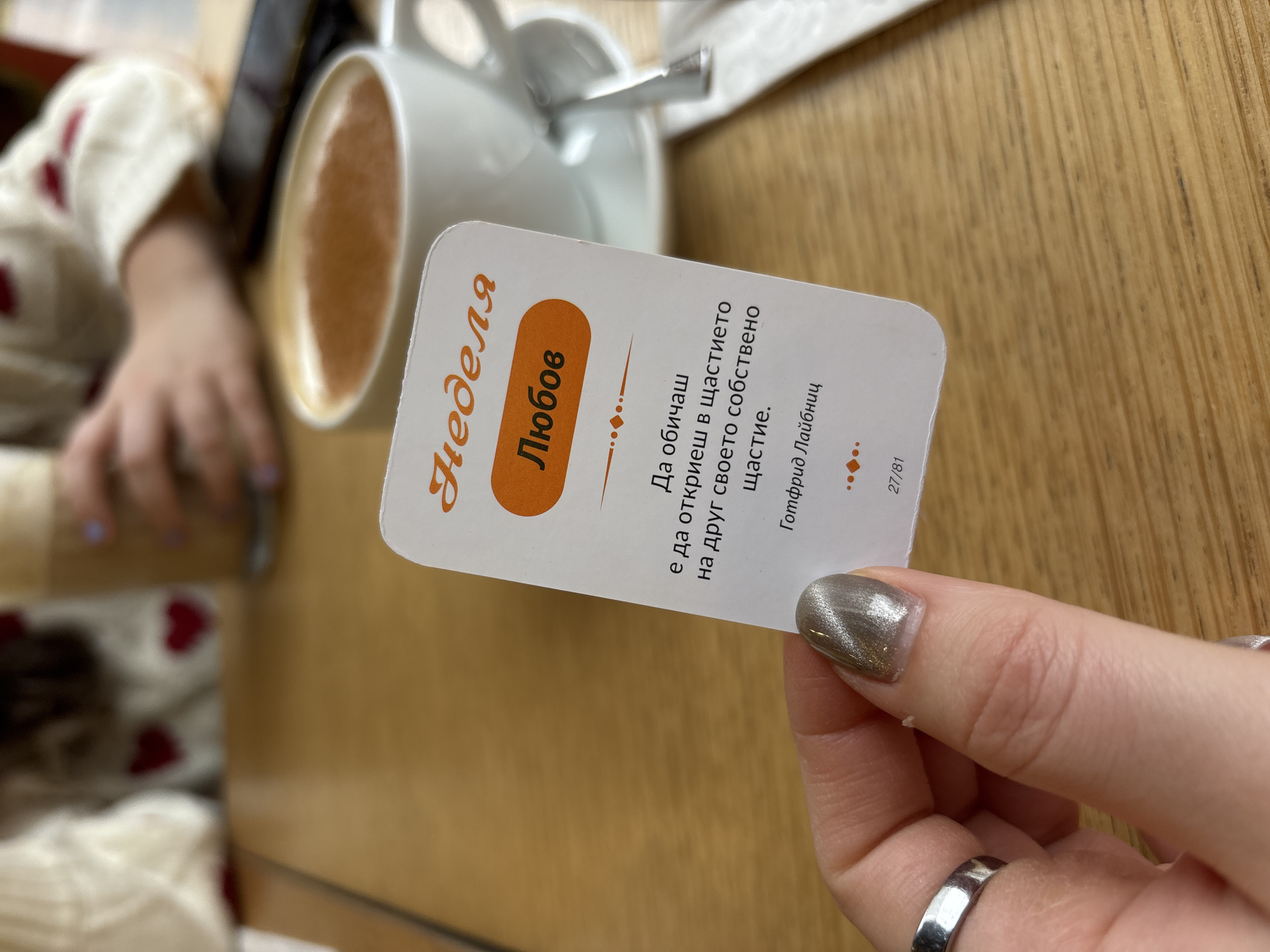 A woman is holding a card with the text "Неделя Любов" (Sunday Love) in Bulgarian. The card has an orange rectangle and text on it. The text is a quote by Gottfried Leibniz that translates to "To love is to find in the happiness of another your own happiness." The card also has the number "27/81" printed on it. A cup of coffee sits on the table behind the card. The woman is wearing a ring on her finger. The table is made of wood. The photo was taken at a table at a cafe in Varna, Bulgaria.