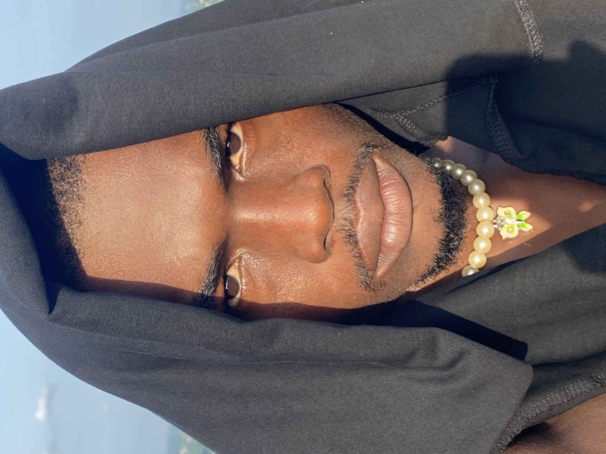A close-up, eye-level, medium shot captures a dark-skinned person wearing a black hood and a pearl necklace with a butterfly pendant. The person is looking directly at the camera, with their face partially obscured by the hood. The lighting suggests it is daytime, with sunlight casting shadows on the person's face. The background is blurred, showing a bright sky and a hint of landscape, possibly indicating an outdoor setting. The specific location within Abakaliki, Nigeria, cannot be determined from the visual information provided in the image.