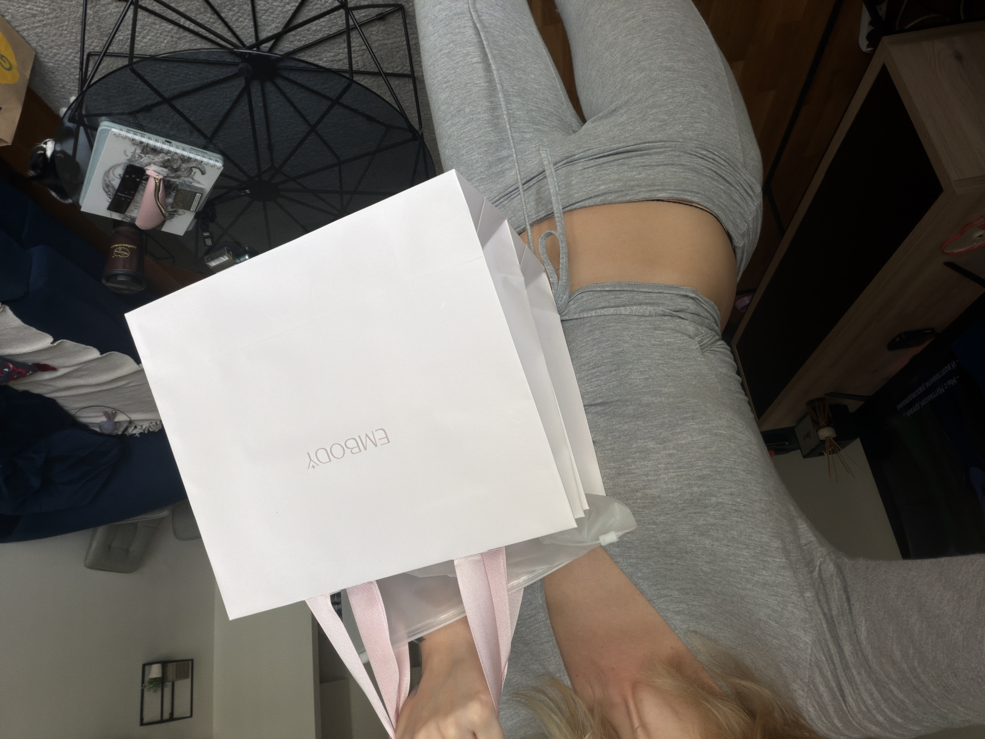 The image depicts a person, likely female, wearing a grey tracksuit. The person is holding a white shopping bag with the word "EMBODY" printed on it in grey. The bag has pink ribbon handles. The person is positioned in front of a black wire-frame table. On the table, there are various items including a notepad with writing on it, and other miscellaneous objects. To the left of the table, a blue couch or section of a couch is visible. In the background, there are parts of a room, including a television, and an indoor houseplant. The setting appears to be indoors, likely a home environment. The location in the city of Belgrade, Serbia cannot be definitively determined from this image.