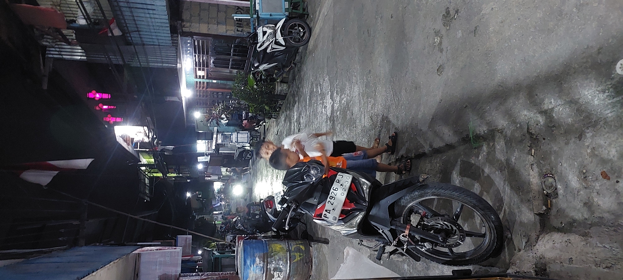 The image depicts a nighttime street scene in what appears to be an urban environment in Jayapura, Indonesia. Two young boys are the primary subjects, standing in the foreground on a paved street. The boy on the left wears an orange shirt and blue shorts, while the boy on the right is in a white t-shirt and dark shorts. They are positioned near a black motorcycle parked on the left side of the street, which has a license plate reading "PA 2926 RJ". The street is illuminated by artificial lights, including some visible from buildings and street lamps. In the background, there are various structures, including buildings with corrugated metal roofs and what appear to be small shops or stalls. A red, white, and blue flag, likely the Indonesian flag, is visible flying from a pole above one of the buildings. Several other motorcycles are parked along the street. The ground is paved and appears somewhat wet, reflecting the light. The overall atmosphere is that of a typical Indonesian street at night, possibly a residential or commercial area.